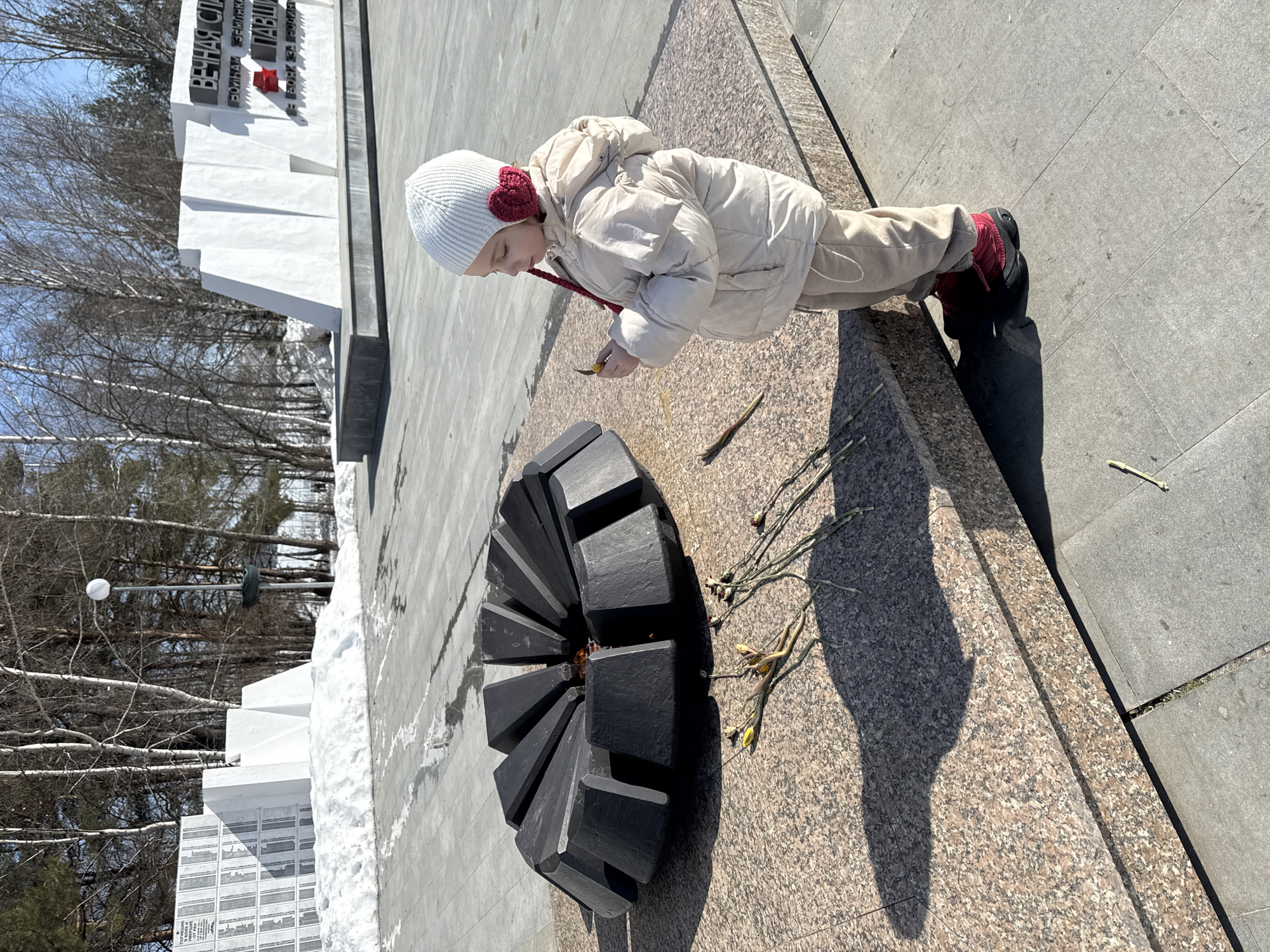 A young child wearing a winter coat, hat, and boots is placing a sprig of greenery near a black, fire-containing memorial. Several other sprigs have already been laid. A white monument with Cyrillic writing is visible behind the memorial. The scene is set in a public outdoor area, likely in the city of Chaikovsky, Russia. The surface is paved with granite and concrete. Bare trees and remnants of snow are visible in the background.