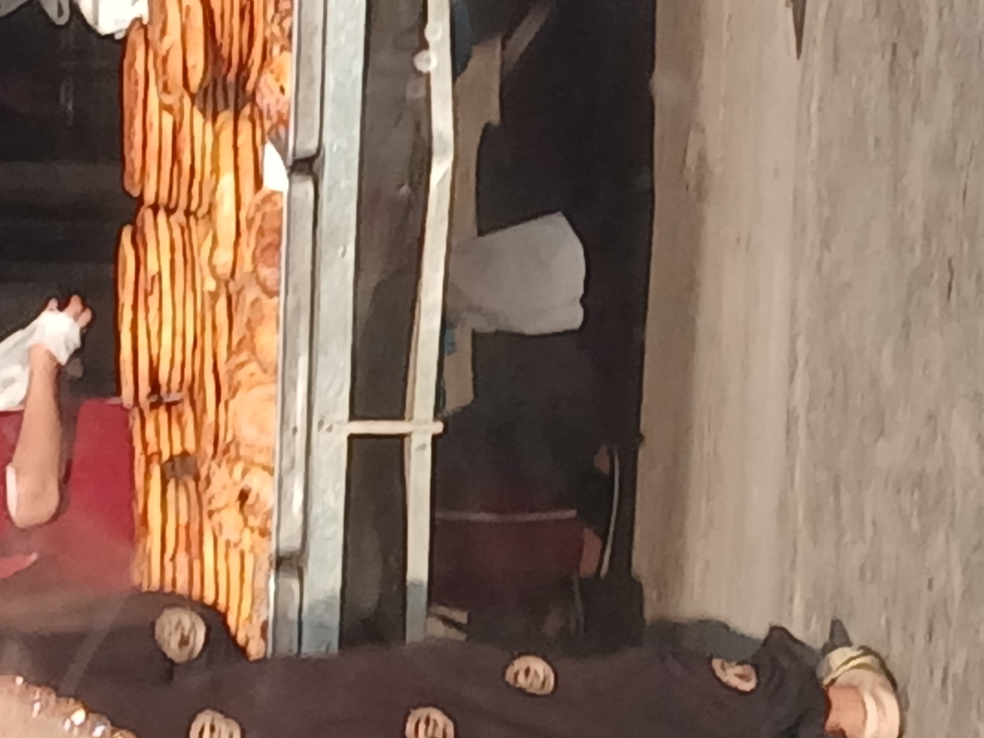 The image depicts a close-up view of stacks of round, flatbreads displayed on metal trays. The bread appears golden brown and is layered in rows. In the foreground, the lower portion of a person's legs and feet is visible. The person is wearing a dark-colored garment with circular patterns and light-colored footwear. A portion of another person's arm and hand, also in a dark garment, is visible on the left side. The background is blurred, but it suggests an outdoor market setting, possibly in Khankiz, Uzbekistan, given the nature of the food display. A white cloth hangs from a rack in the mid-ground. The overall lighting suggests it is daytime.