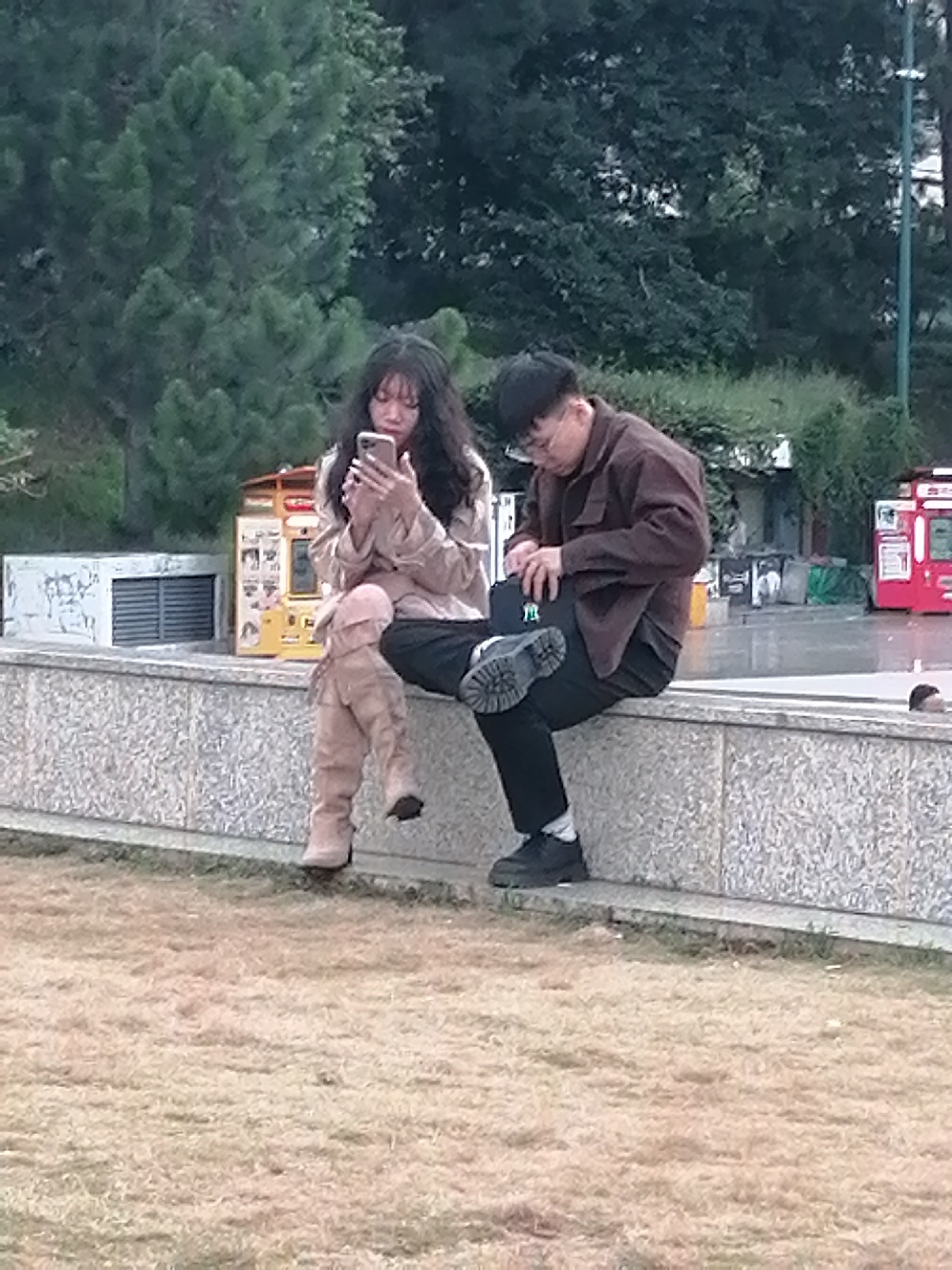 Two individuals, a female and a male, are seated on a stone ledge in what appears to be a park setting. The female, on the left, has long dark hair and is wearing a light brown jacket and thigh-high tan boots. She is holding a smartphone and appears to be looking at it. The male, on the right, has short dark hair and glasses, and is wearing a dark brown jacket and black pants. He is reaching into his lap, possibly retrieving or putting away an item. Behind them, a yellow vending machine is visible, along with a white structure with horizontal slats and a red vending machine in the distance. The ground in front of the ledge is covered in dry grass. The background features lush green trees. Based on the presence of the yellow vending machine, which is commonly found in Da Lat, Vietnam, the location is likely Da Lat.