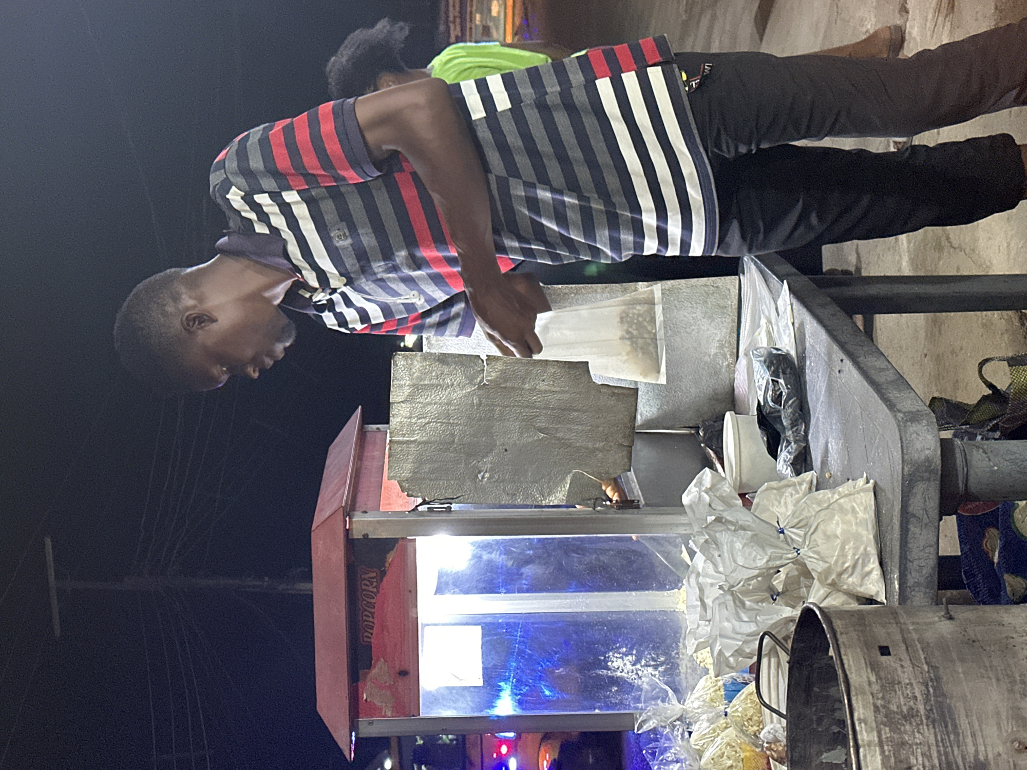 A man is making popcorn at an outdoor stand, with a popcorn machine and filled bags on the counter. He is wearing a striped shirt and dark pants. Another person is standing behind him, wearing a green shirt. The location is a city street at night.