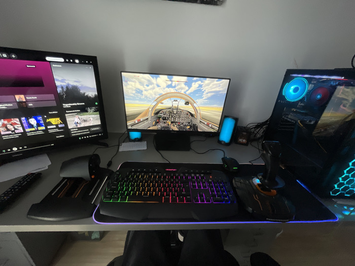 This media file captures a multi-monitor computer setup in what appears to be a home office or gaming room in Libourne, France. The scene is dominated by a desk featuring two monitors, a gaming keyboard, mouse, a desktop PC tower, and specialized flight simulation peripherals.

On the left, a large monitor displays a media player interface, likely Spotify or YouTube Music, with a dark theme and purple accents. Visible elements include album art thumbnails, song titles, and a search bar. The text "Travis Scott" is clearly legible on one section, and "Wojewódzki" is partially visible.

The right monitor shows a first-person perspective from the cockpit of a small aircraft within a flight simulator game. A runway stretches into the distance under a bright, partly cloudy sky, with detailed instrumentation visible inside the cockpit.

Below the monitors, the desk holds various peripherals. A large, black gaming keyboard with vibrant RGB backlighting, featuring a "RAPTOR" brand logo on its spacebar, is centrally placed. To its right, a black gaming mouse with a green LED is positioned on an extended mousepad that also accommodates a flight stick and a throttle quadrant. These flight simulation controllers are black with subtle orange accents. Two cylindrical speakers, emitting a blue light, are situated behind the keyboard and mousepad, flanking the main monitor. A small white mini-keyboard is also visible near the left speaker, next to an object labeled "ice." A remote control rests on the left side of the desk.

The desktop PC tower stands to the far right, showcasing its internals through a transparent side panel. It features internal RGB fans glowing blue and red, and a front panel with a blue honeycomb LED design. Visible wires are present behind the monitors.

The setup suggests the user is engaged in both media consumption and immersive flight simulation gaming. The bright screens and internal lighting of the PC and peripherals create a well-lit environment, though the time of day is not explicitly indicated. No people are visible in the image, but the arrangement implies a single user's dedicated space.