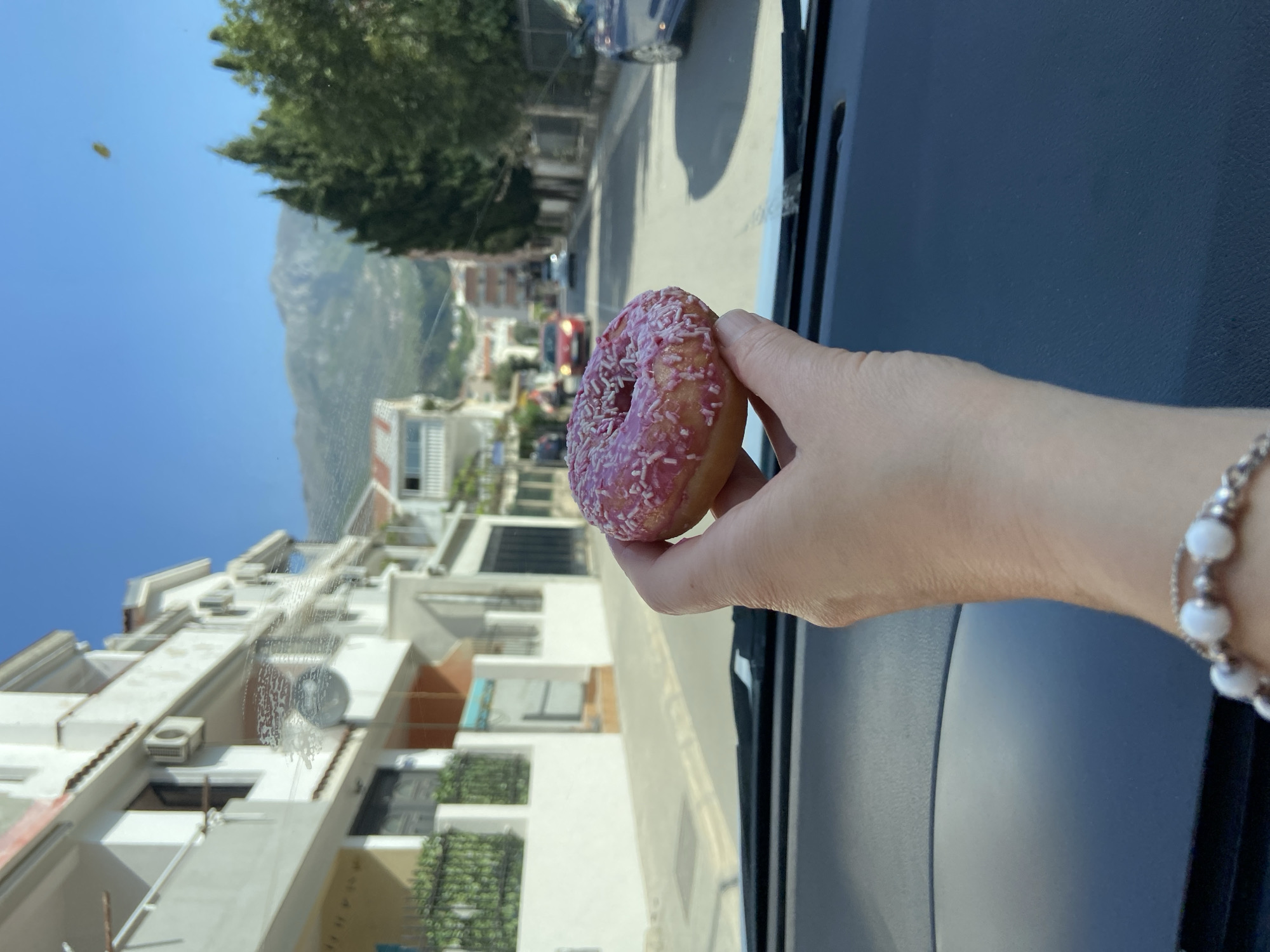 The image, taken from inside a vehicle looking outwards, depicts a casual scene on a bright, sunny day in Budva, Montenegro. In the foreground, a person's hand holds up a single donut. The donut is circular, coated in vibrant pink frosting, and sprinkled with numerous small, lighter-colored candy sprinkles. A silver bracelet adorned with white, spherical beads is visible on the wrist of the hand.
The view through the windshield reveals an urban or semi-urban landscape. Multi-story buildings, predominantly white with visible balconies and exterior air conditioning units, line the street. Green foliage from trees is scattered throughout the scene. In the background, large mountains rise prominently against a clear, azure sky. Several vehicles can be seen on the street, suggesting activity. The bright sunlight indicates it is daytime, likely morning or afternoon. There are some smudges or reflections visible on the windshield. No text is visible in the image.
