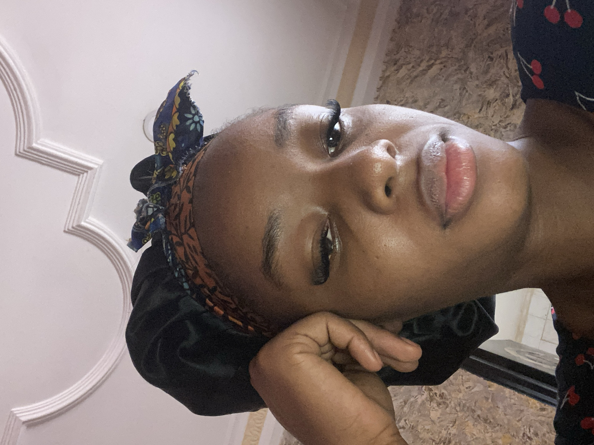 A single individual is depicted in a close-up, low-angle portrait. The individual exhibits a dark skin tone and maintains a frontal gaze towards the viewer. A dark, possibly satin, bonnet covers the upper portion of the head. Below the bonnet, a fabric headband is visible, displaying a pattern of orange and black geometric shapes and a blue section with light blue, yellow, and white floral designs, tied at the top. Both eyes feature prominent, long, dark false eyelashes. The individual's lips are full, glossy, and slightly parted. The left hand is positioned against the left side of the head, with the fingers gently curved. A dark garment adorned with red cherry patterns is visible on the lower right quadrant of the image. The background consists of a white ceiling with an arched, decorative white molding occupying the upper half. The lower left background shows a textured surface with mottled brown and beige tones. No visual identifiers within the image provide information to confirm the location as Jos, Nigeria.