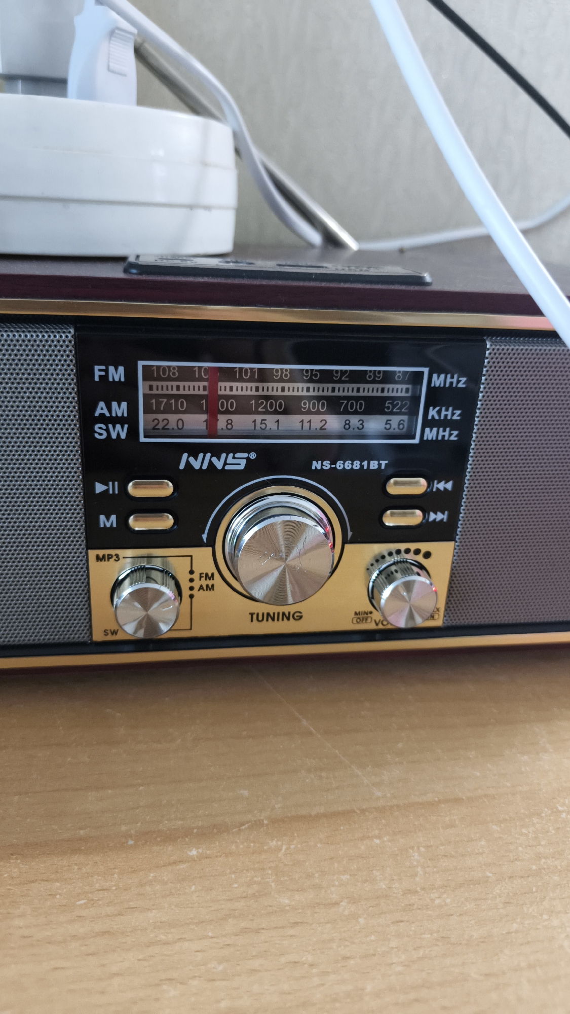 The image shows a close-up, slightly angled view of a portable radio, likely a vintage or retro style model, resting on a wooden surface. The radio is predominantly black with gold accents and features a display screen showing frequency bands (FM, AM, SW) and numerical values. There are several prominent control knobs: a large tuning knob in the center, a smaller volume/power knob on the right, and another smaller knob on the left likely for band selection or mode. Button controls for playback and skipping tracks are also visible. The radio's brand name "NNS" and model number "NS-6681BT" are printed on the front. In the background, partially out of focus, are white and grey electrical cords and a white electrical plug, suggesting the radio is either plugged in or charging. The wooden surface appears to be a table or desk, with visible grain and some minor scuff marks. The overall lighting suggests an indoor environment, possibly a home or office. There is no indication of the city of Vladimir, Russia, or any other specific geographic location within the image itself.