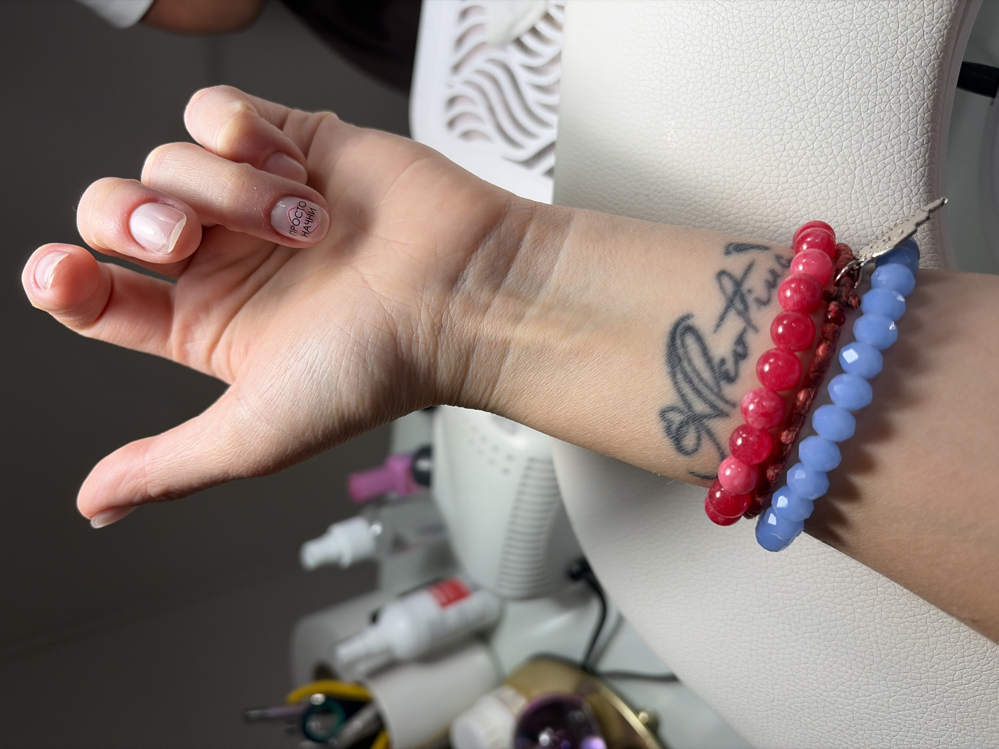A photograph shows a person's hand and wrist wearing three beaded bracelets (red, light blue, and brown) on a light skin tone. The hand is extended with fingers slightly curled. The fingernails appear to have a clear or light pink coating; one nail has letters on it. A tattoo is visible on the wrist near the bracelets. The background shows parts of a white salon chair or workstation, various bottles (likely nail products), and other equipment. The scene suggests a nail salon or similar beauty establishment. The image does not directly indicate the location of Budva, Montenegro; this association is not apparent.