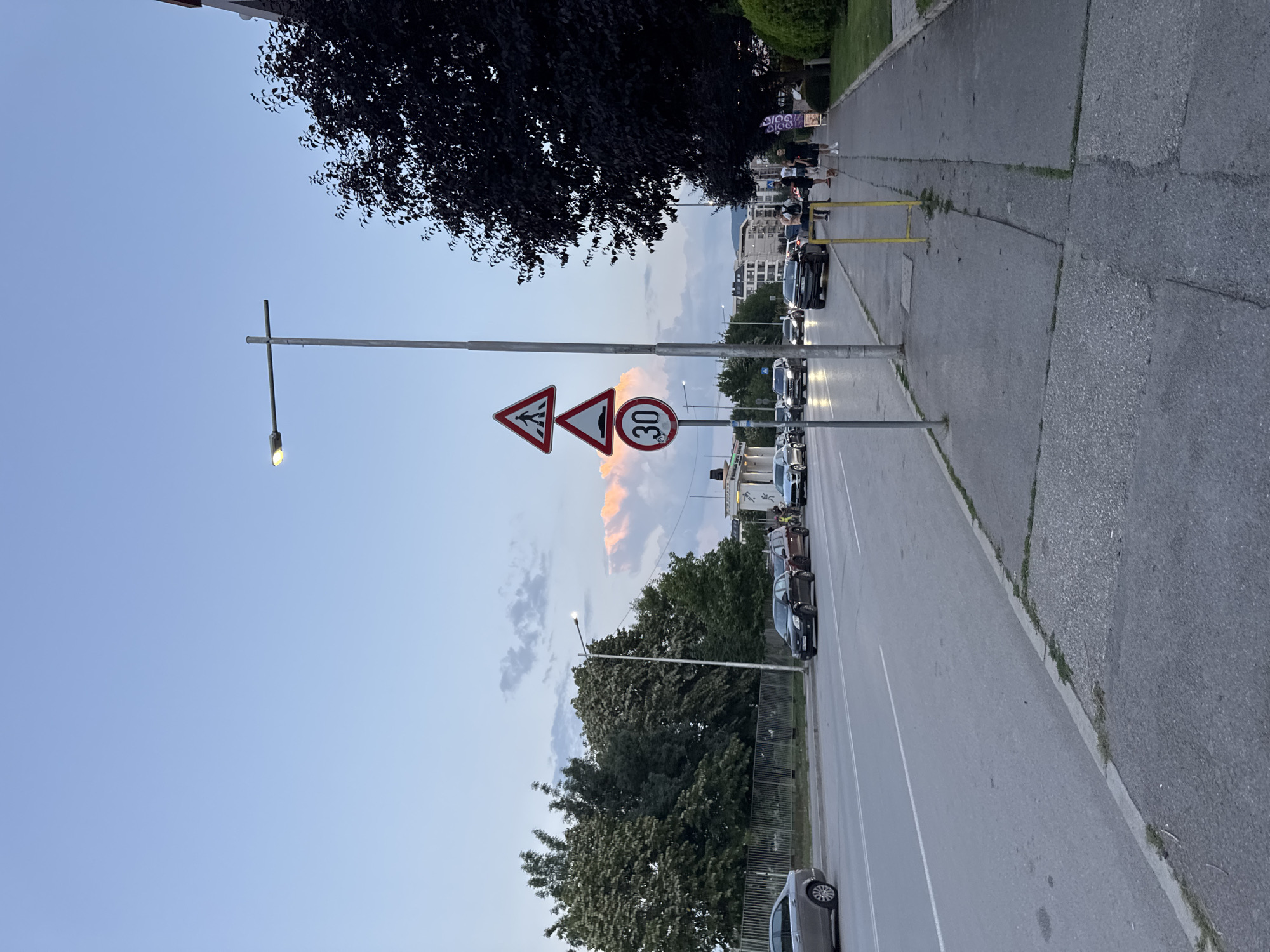 The image depicts a street scene in Sofia, Bulgaria, at dusk. A multi-lane road runs horizontally across the frame, with several cars visible. On the right side of the road, a sidewalk is lined with pedestrians and a dark leafy tree. A tall street lamp with a single illuminated bulb stands on the sidewalk, accompanied by three traffic signs: a pedestrian crossing warning, a speed bump warning, and a circular sign indicating a speed limit of 30. The sky above is a soft blue with hints of pink and orange from the setting sun, illuminating a distant cloud formation. The overall environment appears to be an urban area with buildings visible in the background.