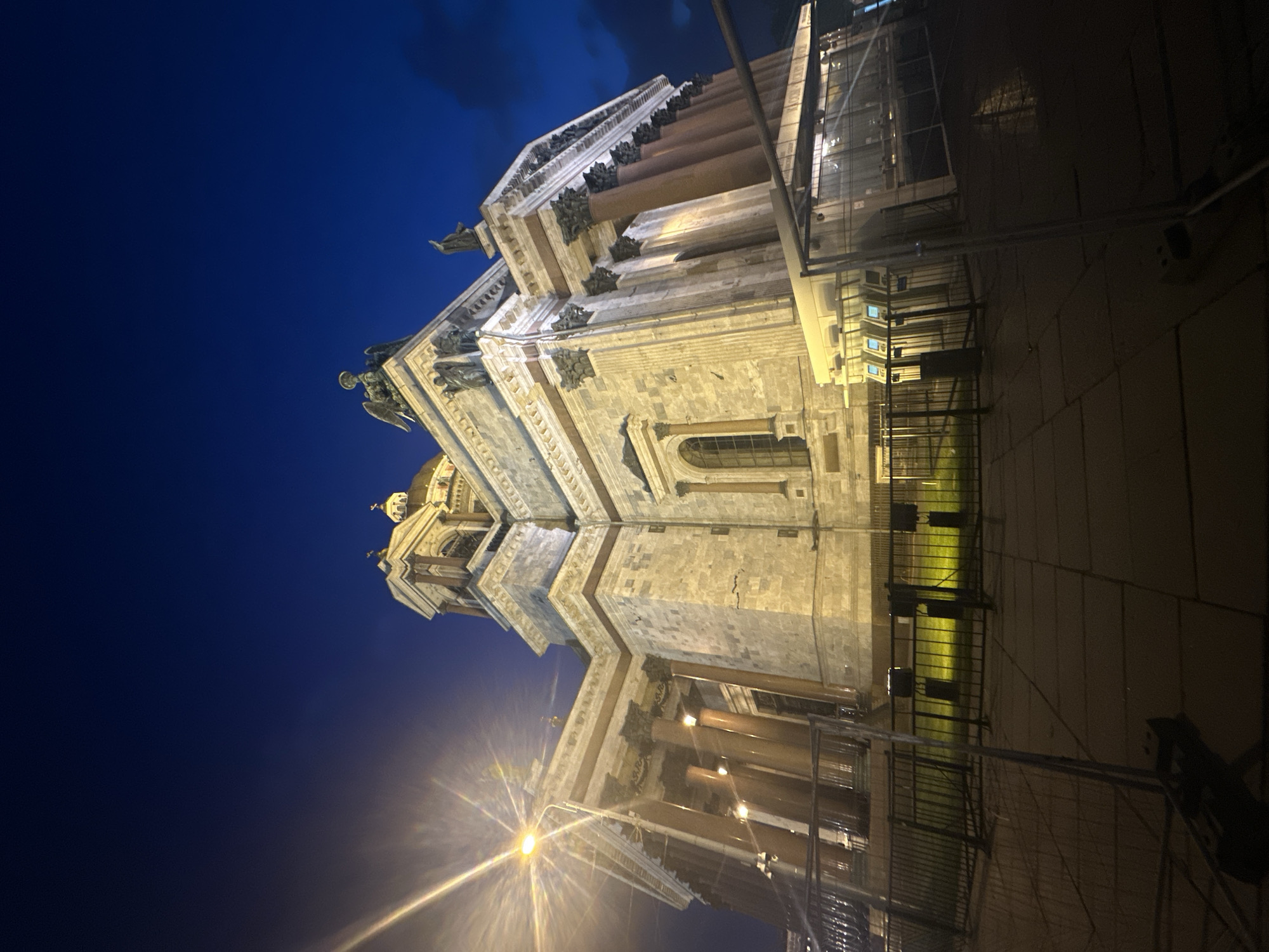 The image captures a low-angle view of St. Isaac's Cathedral in Saint Petersburg, Russia, at night. The cathedral is brightly lit, accentuating its classical architectural details like columns, statues, and the dome. The sky is a deep blue, contrasting with the illuminated facade of the cathedral. Scaffolding and a temporary structure are visible to the right of the cathedral, suggesting ongoing maintenance or construction work. A street light casts a bright flare at the bottom of the image.