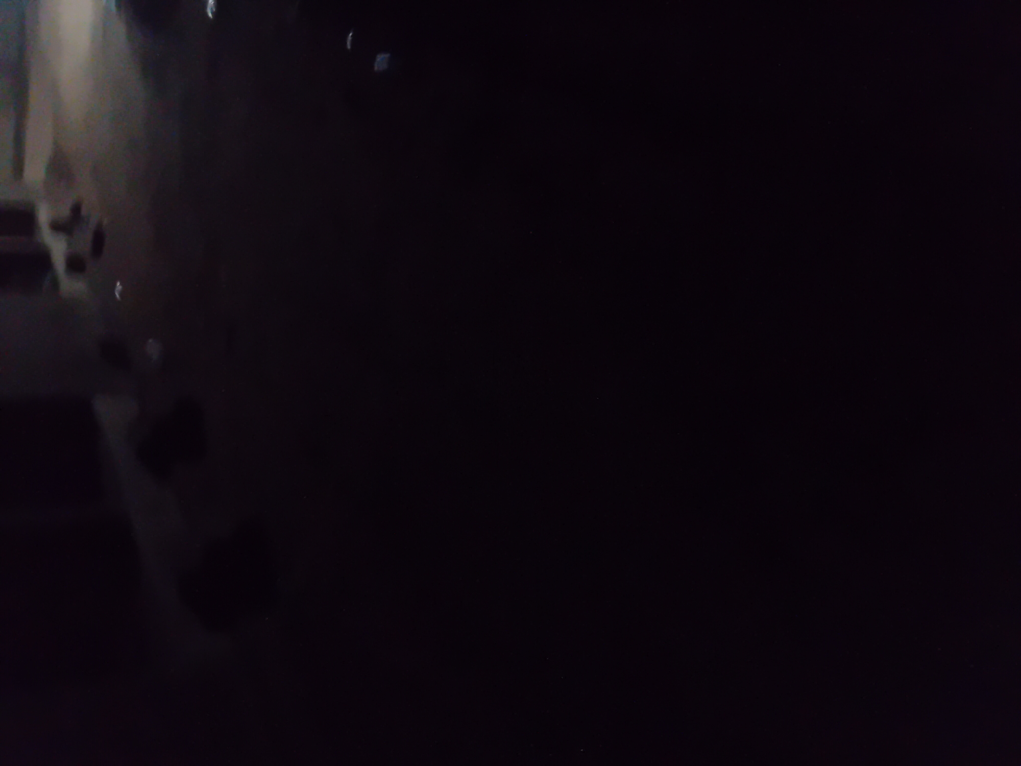 The image shows a dark, blurry scene at night, likely in an outdoor environment. In the upper portion of the image, some indistinct shapes suggest buildings or structures. There are also lighter patches that could be reflections or distant lights. The foreground is almost entirely black, making it impossible to discern any specific details of the ground or any objects or people present. The overall impression is one of low light conditions and limited visibility. The provided location of Jauro Yinu, Nigeria, offers a geographical context, but the image itself provides no direct visual evidence to confirm this location.