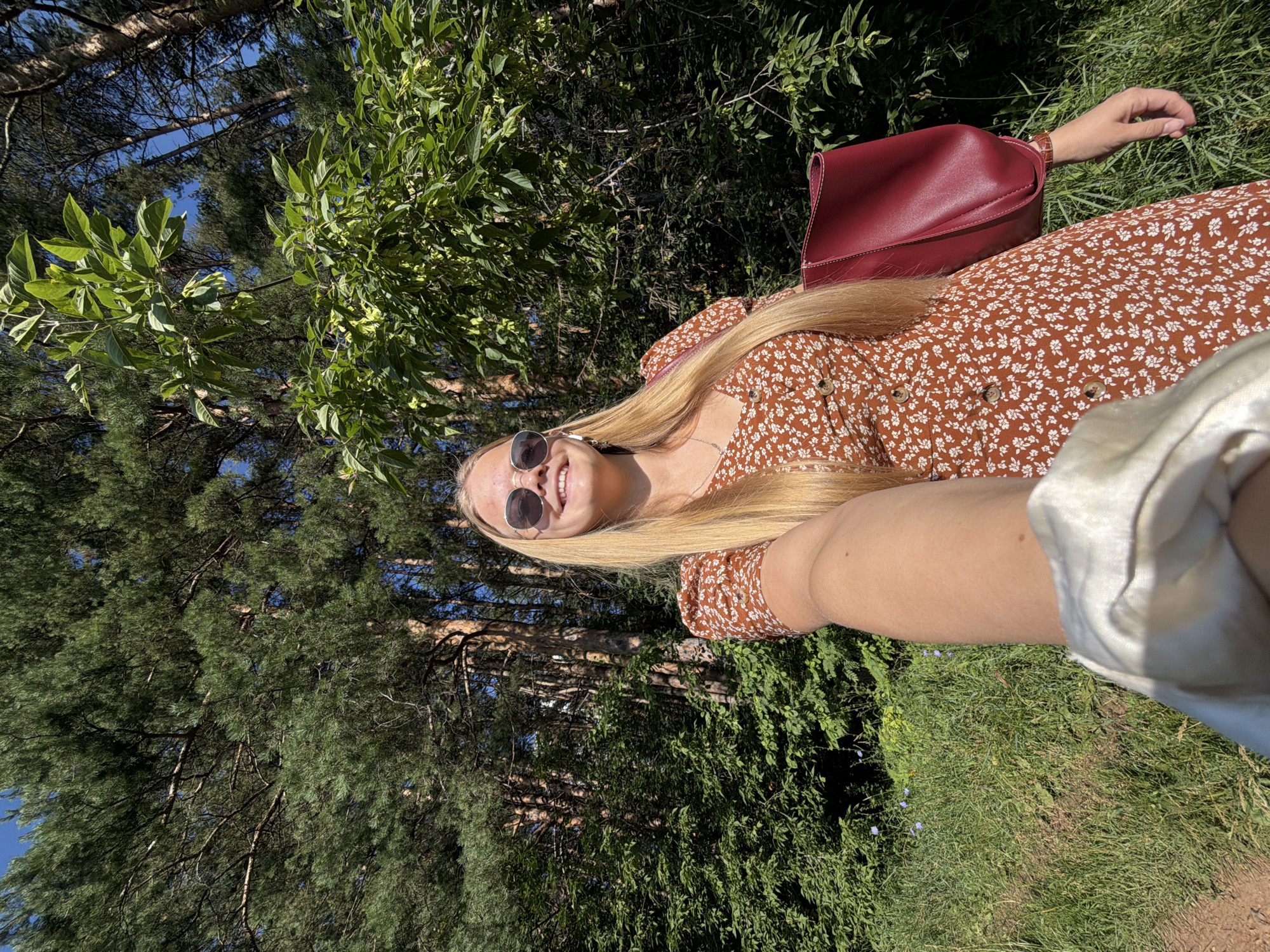 A young woman with long blonde hair and sunglasses is taking a selfie outdoors. She is wearing a brown floral dress and holding a red purse. She is standing in a grassy area with trees in the background. The image appears to be taken on a sunny day. The location is the Izhevsk zoo, Russia, a popular spot for locals and tourists alike. The woman is smiling and looking at the camera. She has a light tan and appears to be in her early twenties. The trees are tall and lush, and the grass is green and well-maintained. The zoo is home to a variety of animals, and it is likely that the woman is visiting with friends or family. The zoo is also a popular place for outdoor events, and it is possible that the woman is attending a concert or festival.