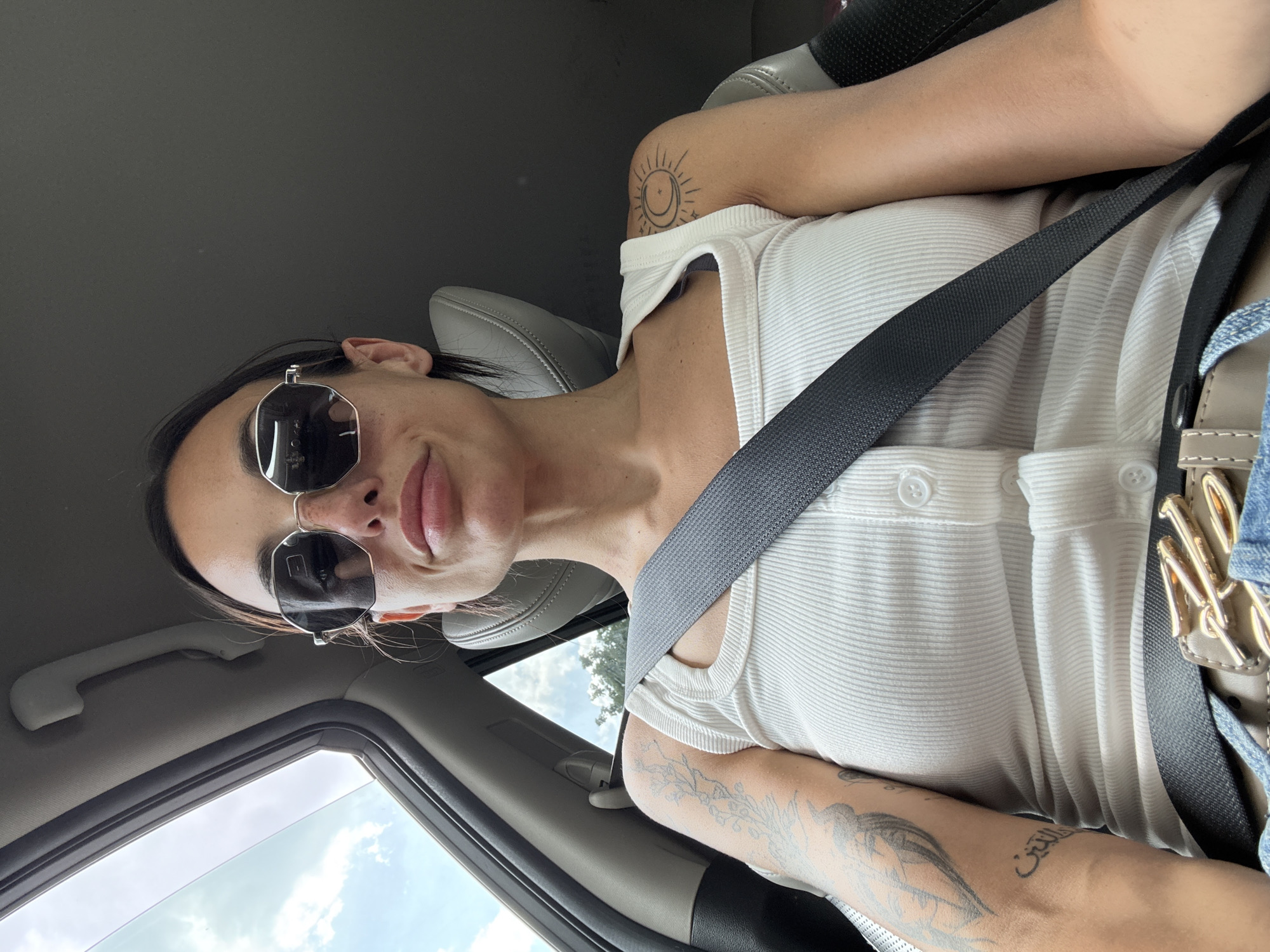 A woman with dark hair is seated in the passenger seat of a car, wearing sunglasses, a white ribbed tank top, and a grey seatbelt. She has tattoos on her left arm and right shoulder, depicting floral designs, script, a sun with a moon and stars, and a bird. The interior of the car is visible, with a sunroof open to the sky. The image was taken in the city of Pleven, Bulgaria.