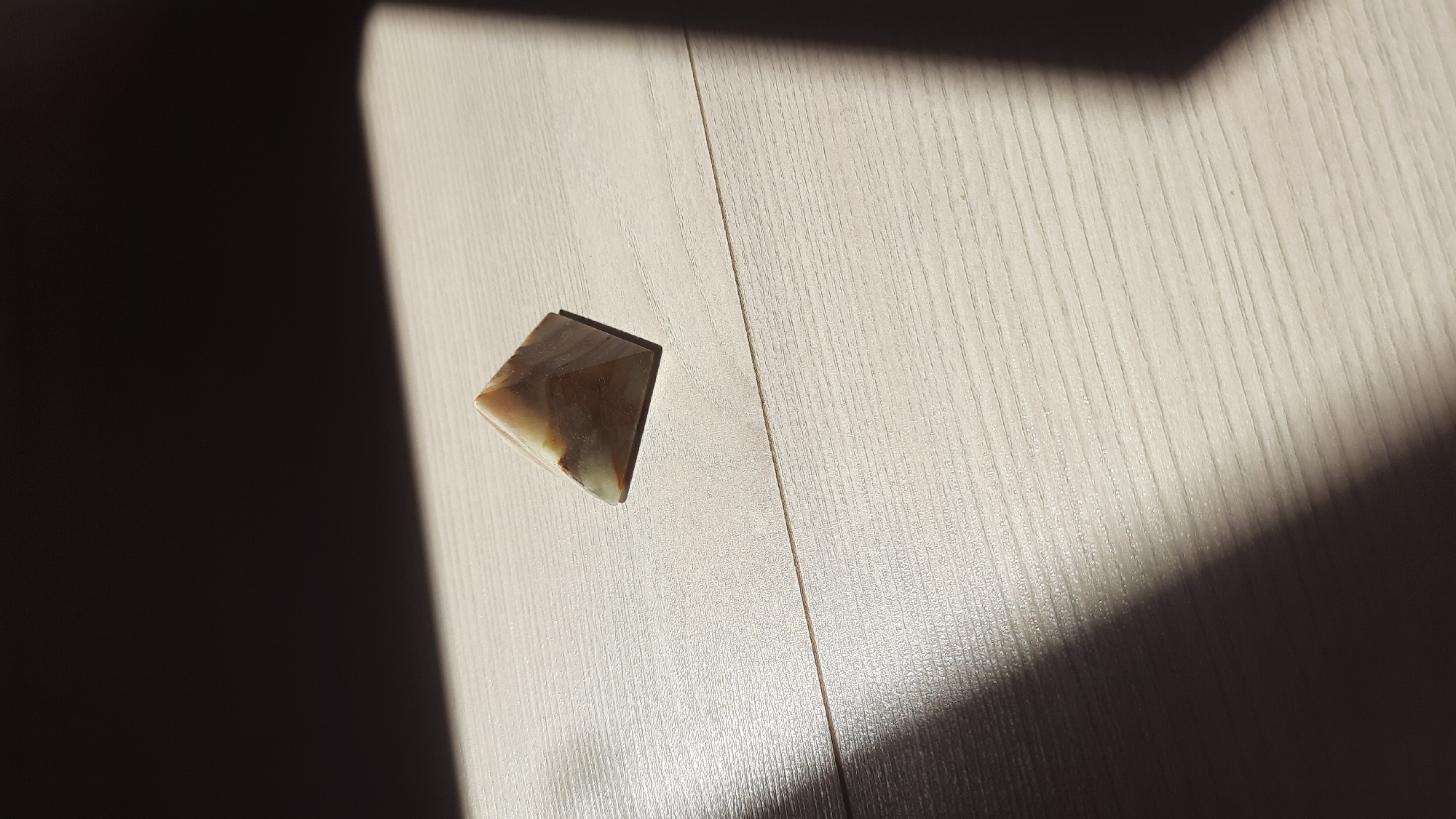 A small, geometric pyramid made of translucent, marbled stone sits on a light-colored wooden surface. Sunlight casts a sharp shadow across the surface, obscuring a portion of the pyramid and the background. The wood grain is visible, oriented horizontally across the frame. The scene is devoid of human presence, and there are no discernible landmarks to indicate a specific location within Desnogorsk, Russia. The image is a still, close-up shot of the object and its immediate environment.