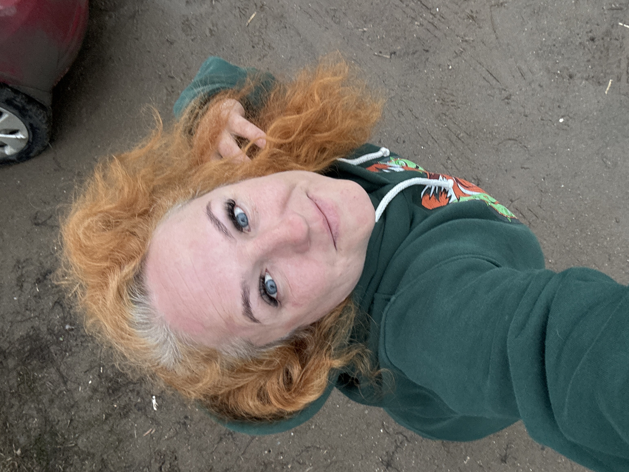 A light-skinned woman with red, curly hair and blue eyes is looking up at the camera. She is wearing a dark green hooded sweatshirt. The background appears to be a ground surface. The image was taken outside. There is no indication of this image being from Chaikovsky, Russia. A red car is partially visible on the left.