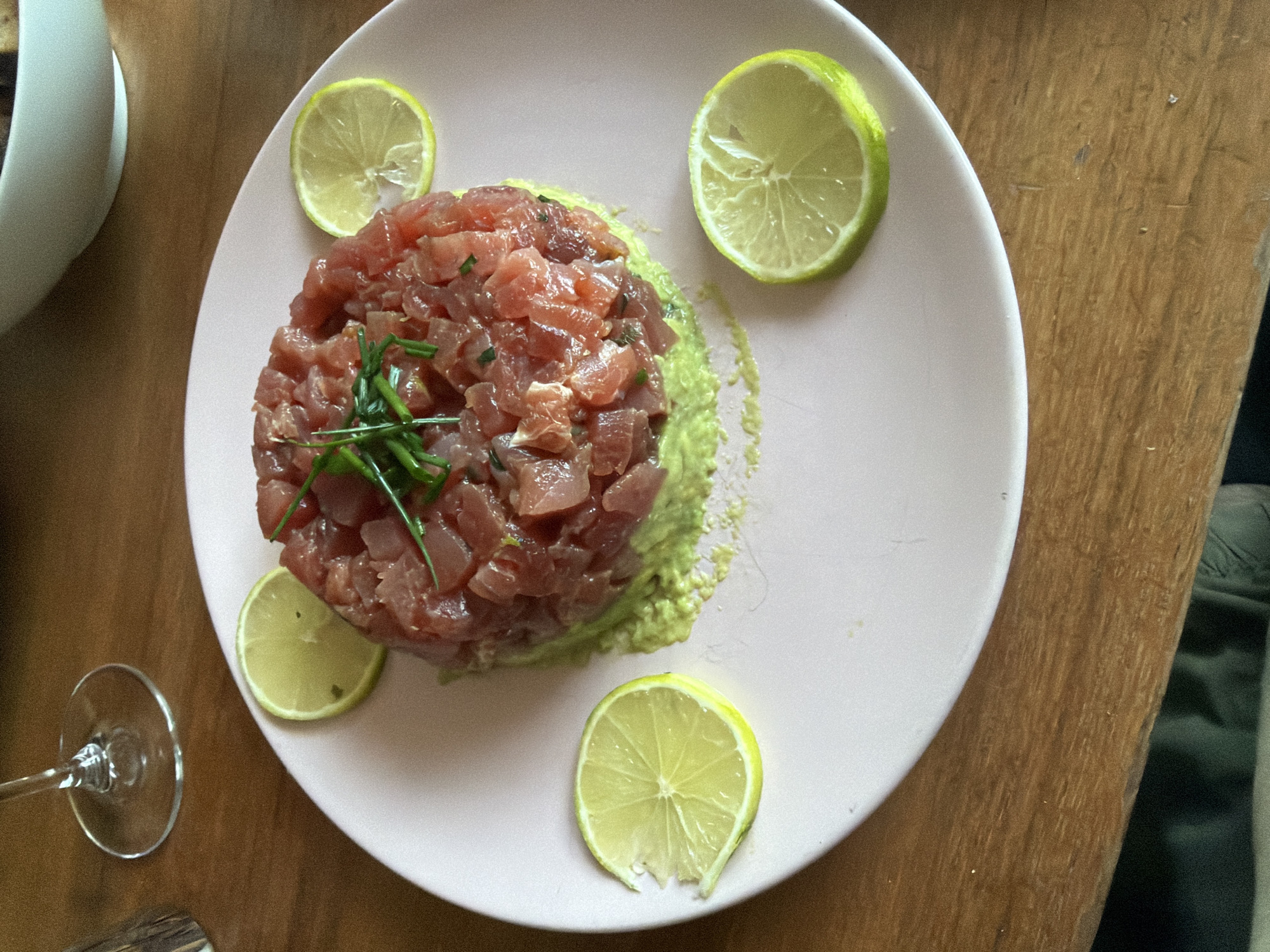 A plate of tuna tartare is centrally positioned on a wooden table, possibly indoors. The tartare is finely diced and sits atop a bed of avocado. The plate is decorated with four lime wedges. A wine glass and a white bowl are partially visible to the left of the plate. Clothing is present in the bottom right corner. The location appears to be a restaurant. The city of London, United Kingdom, cannot be verified from the image.