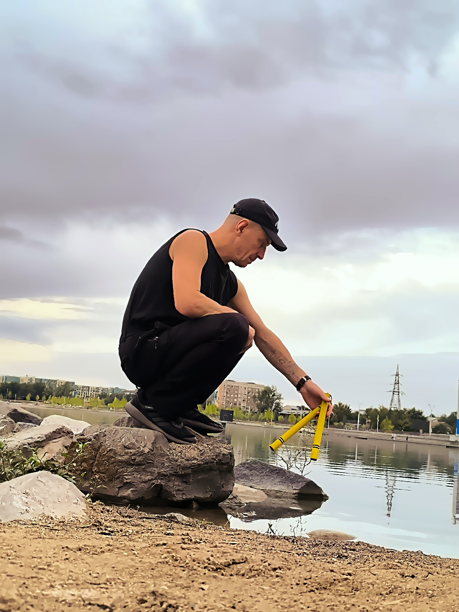 In Almaty, Kazakhstan, a man in black clothing and a black baseball cap crouches on rocks by a body of water. He is holding a pair of yellow nunchaku, seemingly about to dip them into the water. The background shows a cityscape with buildings and trees, under a cloudy sky. The foreground is a sandy bank with more rocks and sparse vegetation.