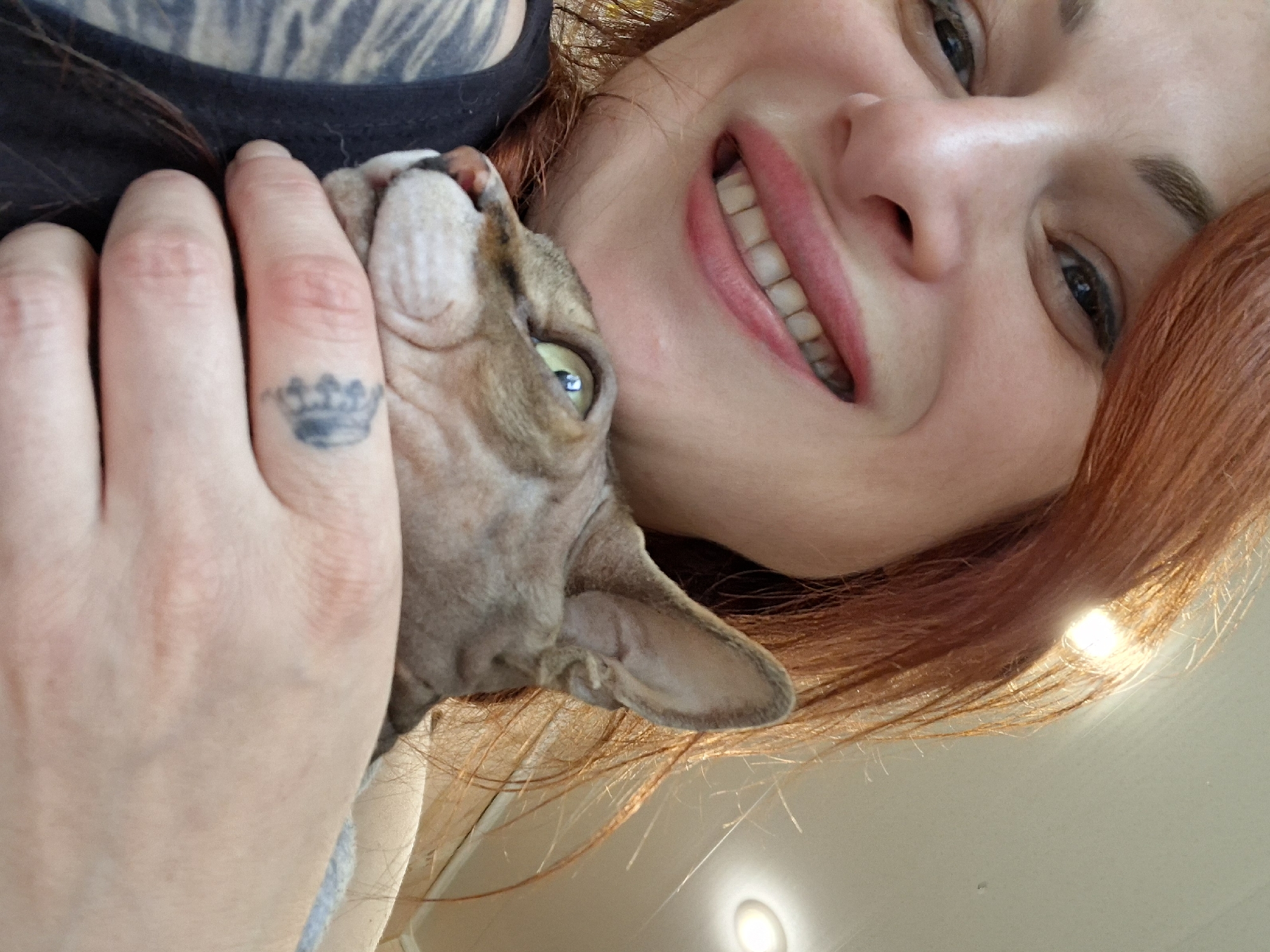 A close-up, selfie-style photograph shows a woman with auburn hair smiling and holding a Sphynx cat. The woman is wearing a black tank top and has tattoos visible on her arm. She has light skin and is wearing lipstick. The cat is light-skinned and appears to be looking away from the camera. The background is out of focus and appears to be indoors with lighting fixtures visible on the ceiling. There is a small tattoo of a crown on the woman's finger. The image was taken in Myadzyel, Belarus.