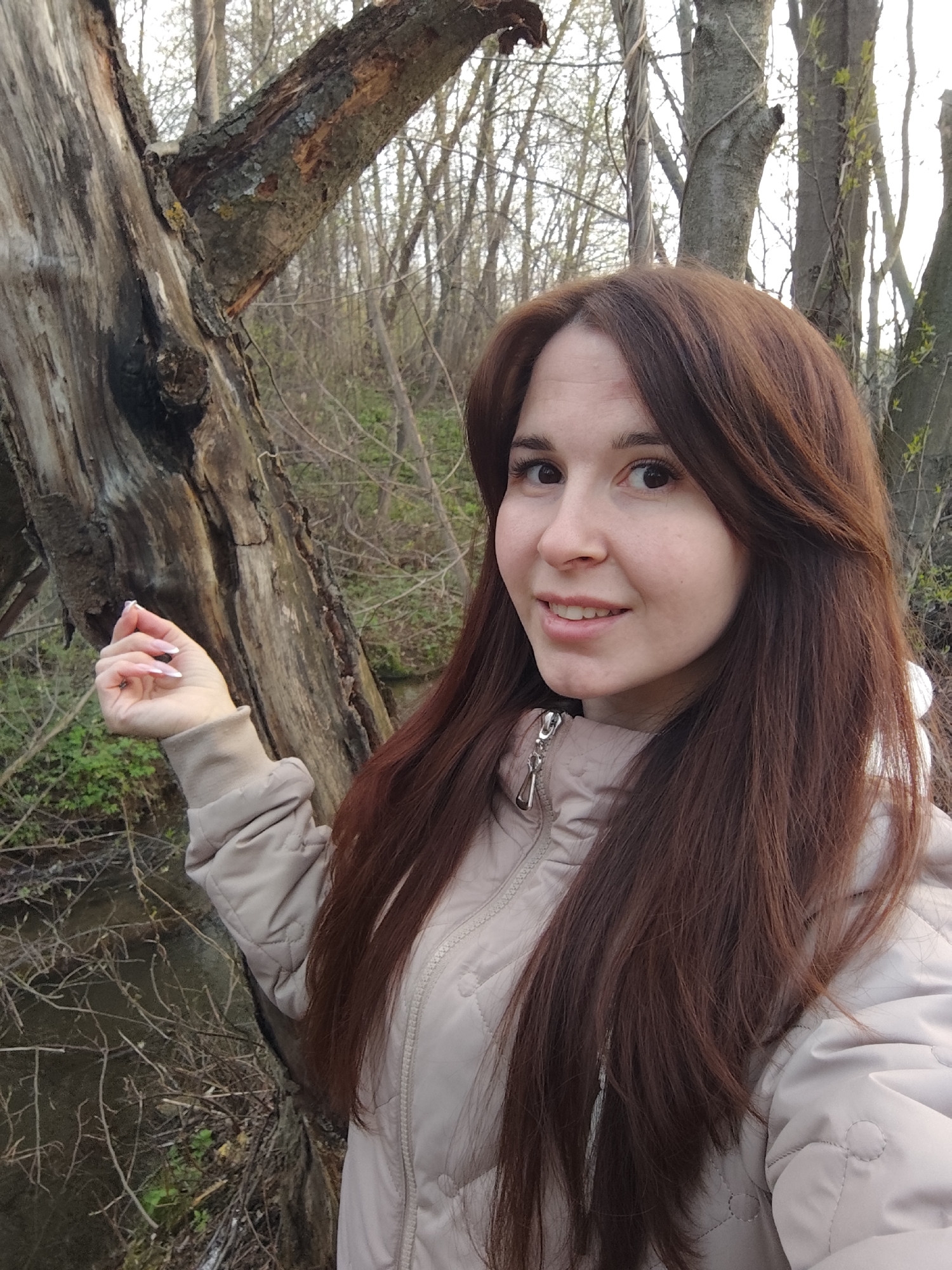 A young woman with long brown hair stands next to a decaying tree in a wooded area. She is wearing a beige jacket. Behind her, a small stream or ditch is visible, along with other trees in the background. The photo appears to have been taken in Фоки, Russia.