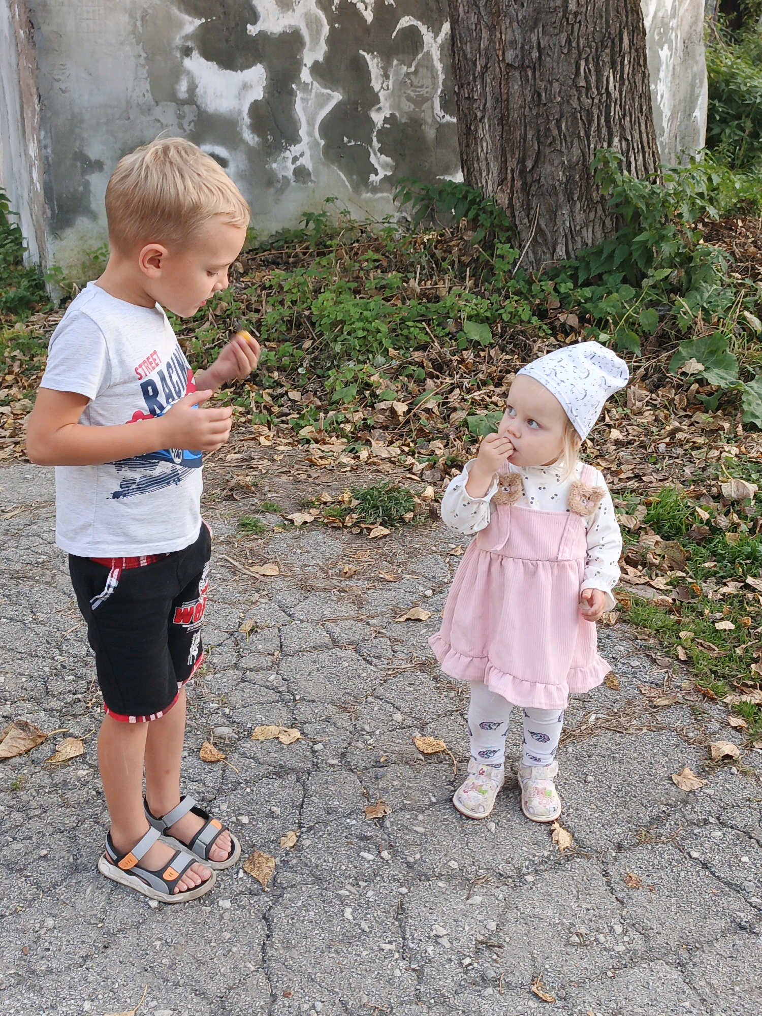 Two young children are standing on a cracked asphalt path in an outdoor environment. The child on the left is a blonde boy wearing a white t-shirt with a "Street Racing" graphic, black shorts with red trim, and grey and orange sandals. He is holding a small orange object in his right hand and appears to be looking at it. The child on the right is a young girl wearing a white long-sleeved shirt with a moon and stars pattern, a pink corduroy overall dress with ruffled hem, white leggings with feather patterns, and light-colored shoes. She is looking up and has her fingers near her mouth. Behind the children, there is a large tree trunk with rough bark, green foliage, and fallen dry leaves on the ground. A weathered concrete wall with abstract dark marks is visible to the left of the tree. The scene suggests an outdoor recreational area or park. It is not possible to determine the precise location within Zlatoust, Russia, from the visual information provided.
