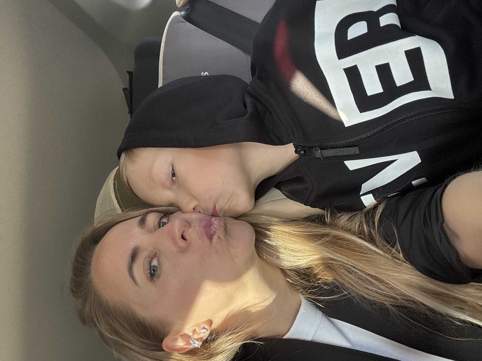 In a close-up, over-the-shoulder shot taken from inside a vehicle, a woman with blonde hair and blue eyes puckers her lips for a kiss towards a young boy. The woman is wearing a black blazer over a white ribbed top and a prominent silver earring with cubic zirconia stones. The boy, with light brown hair, wears a black hooded jacket with a large white "VER" logo on the chest. The background shows car interior elements, including a grey headrest and seatbelt, hinting at a journey within Chaikovsky, Russia. The sunlight casts a warm glow, illuminating their faces and hair.