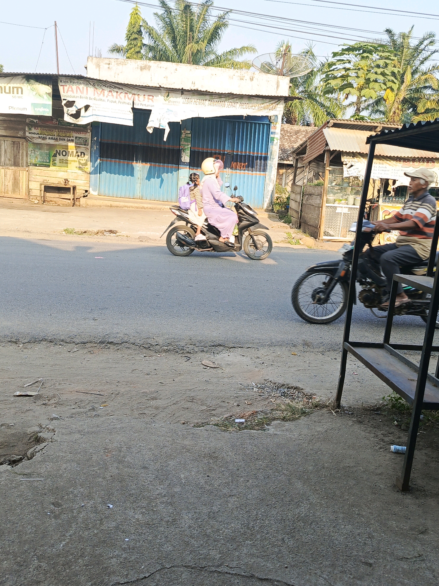 The image depicts a street scene in Megang Sakti I, Indonesia, with a focus on two individuals on motorcycles. On the left, a person wearing a light purple tunic and a yellow helmet rides a black motorcycle, carrying another person on the back who is also wearing a purple outfit and a backpack. They are moving away from the viewer. On the right, an older man wearing a striped polo shirt and a beige cap rides a black motorcycle, also moving away from the viewer. He appears to be looking towards the left.
The environment includes a paved road with some debris and uneven surfaces. Along the side of the road, there are several small shops with corrugated metal roofs and wooden stalls. Banners and signs are visible on the shops, some in Indonesian. Notably, one banner reads "TANI MAKMUR," which translates to "Prosperous Farmer." Palm trees and other tropical vegetation are present in the background, suggesting a warm climate. The lighting indicates it is daytime, likely late afternoon due to the long shadows. A satellite dish is mounted on a building in the background. In the foreground, a metal shelving unit is partially visible on the right.