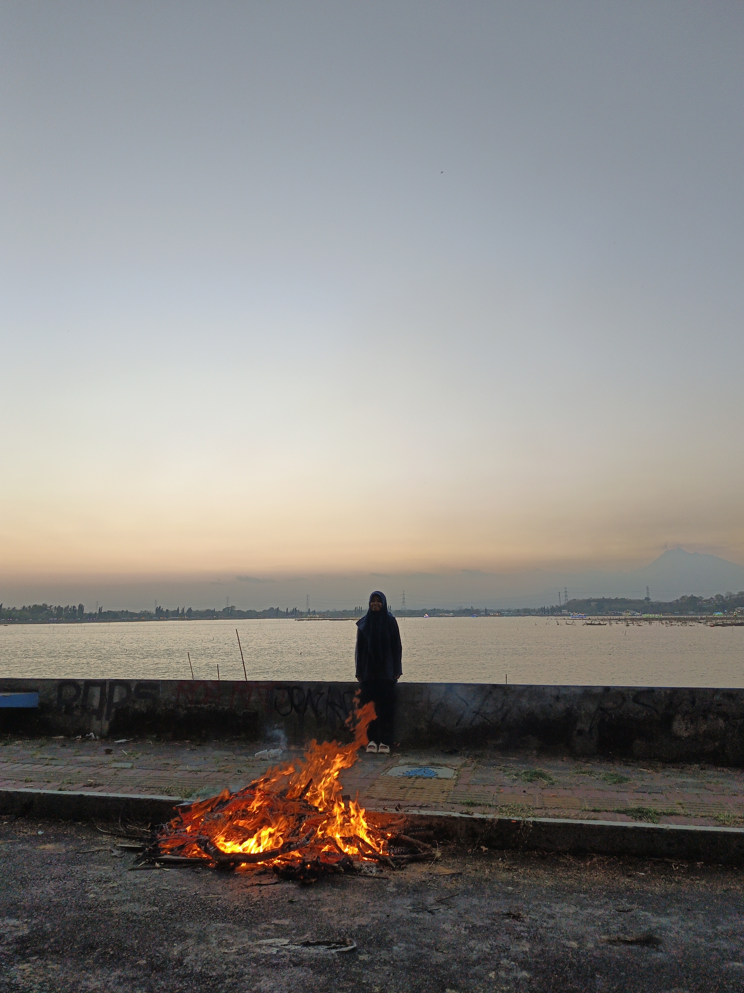 A lone person wearing a hijab stands facing the viewer in front of a calm body of water at dusk. To their left, a large bonfire burns on a paved area near a concrete wall. The sky is a gradient of light blue and soft orange, indicating the sun has recently set. In the distance, a hazy shoreline with trees and structures is visible, and a faint outline of a mountain can be seen on the right horizon. The ground is dark asphalt with some scattered debris.
