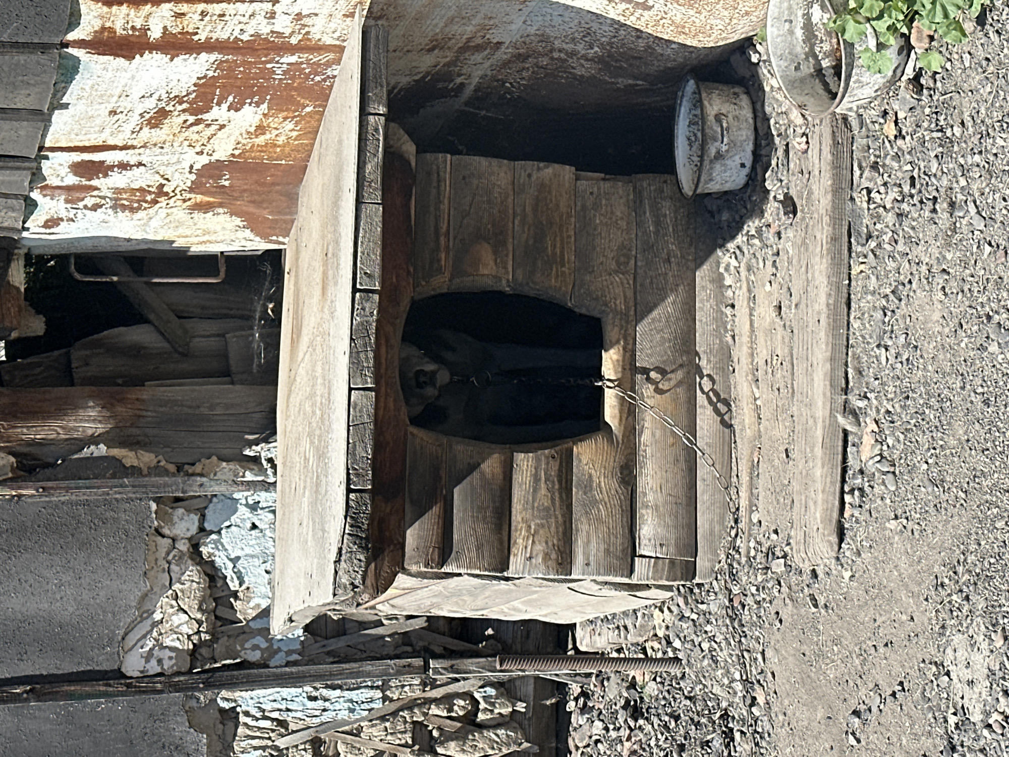 The image shows a close-up, overhead view of a rustic, weathered wooden doghouse set in a gravelly, outdoor setting. The doghouse is aged and shows signs of disrepair, with uneven wooden planks forming its structure. A dark, arched opening serves as the entrance, and a metal chain is attached to the side, suggesting a tether.
Inside the doghouse, a dark-furred dog is visible, partially obscured by the shadows and the structure of the entrance. The dog appears to be lying down or resting. The overall scene conveys a sense of neglect and poverty, with discarded metal containers and debris scattered around the doghouse.
The background is comprised of rough ground covered in gravel and some dirt. To the left, there are crumbling stone and concrete structures, possibly remnants of an old building or foundation, also showing significant weathering. A rusted metal sheet with peeling paint is visible above the doghouse, adding to the dilapidated aesthetic.
The lighting suggests it is daytime, with harsh shadows indicating a bright sun. There are no discernible people in the frame. The location context of Novoyeniseyka, Russia, implies a potentially rural or remote environment where such a humble dwelling for a dog might be found. No text is visible in the image. The overall mood is somber and a little melancholic, focusing on the animal's simple existence in a harsh environment.