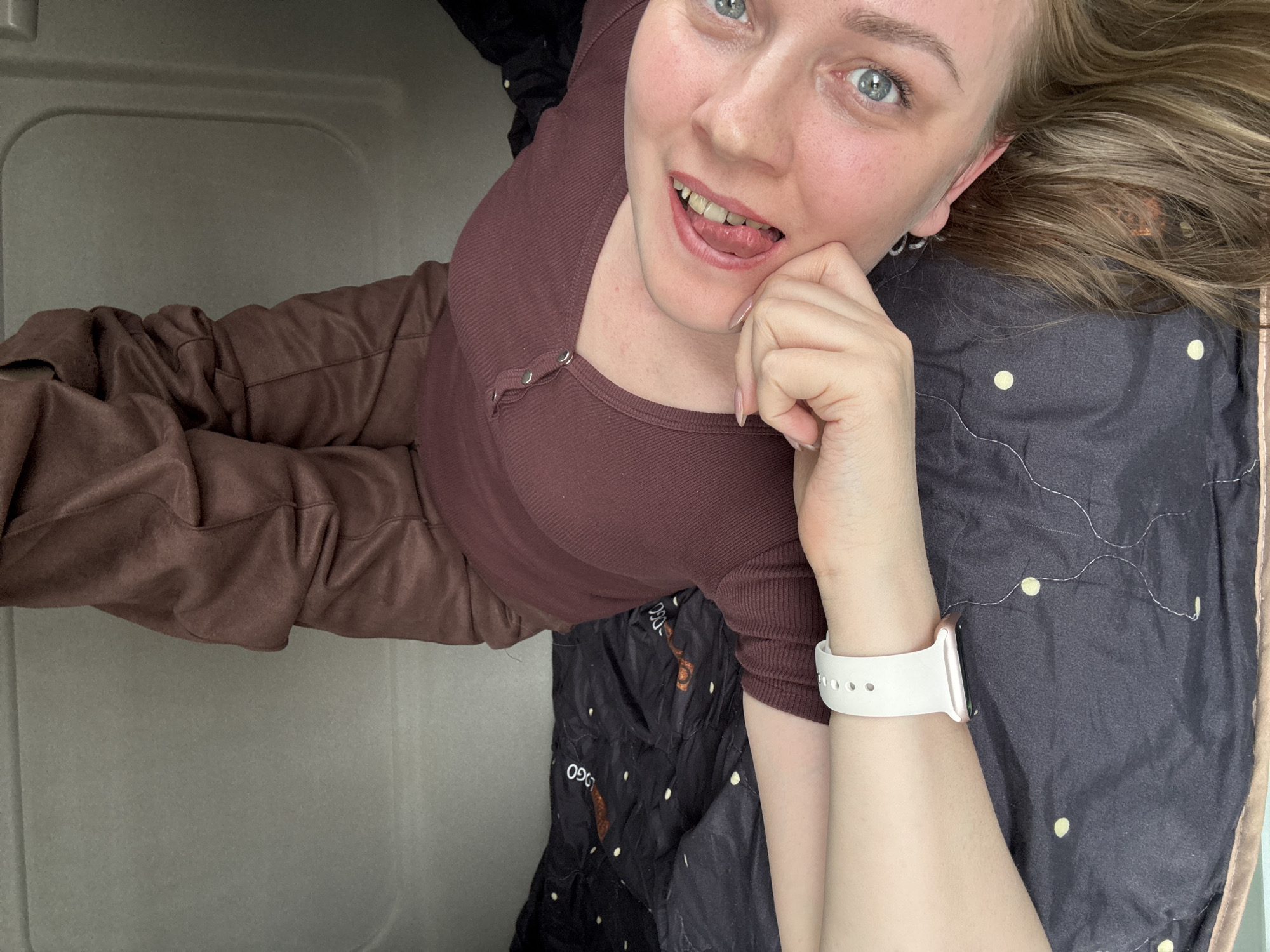 A fair-skinned woman with blonde hair and blue eyes is lying down. She is wearing a dark red-brown ribbed top and dark brown trousers. She is wearing a white smartwatch on her left wrist and a silver hoop earring in her left ear. Her left hand is raised to her face. She is sticking her tongue out. She is lying on a grey quilted blanket with white and brown decorative images. She is in an enclosed, white, capsule-like space with curved edges. The location in Izhevsk, Russia, is not discernible from the image.
