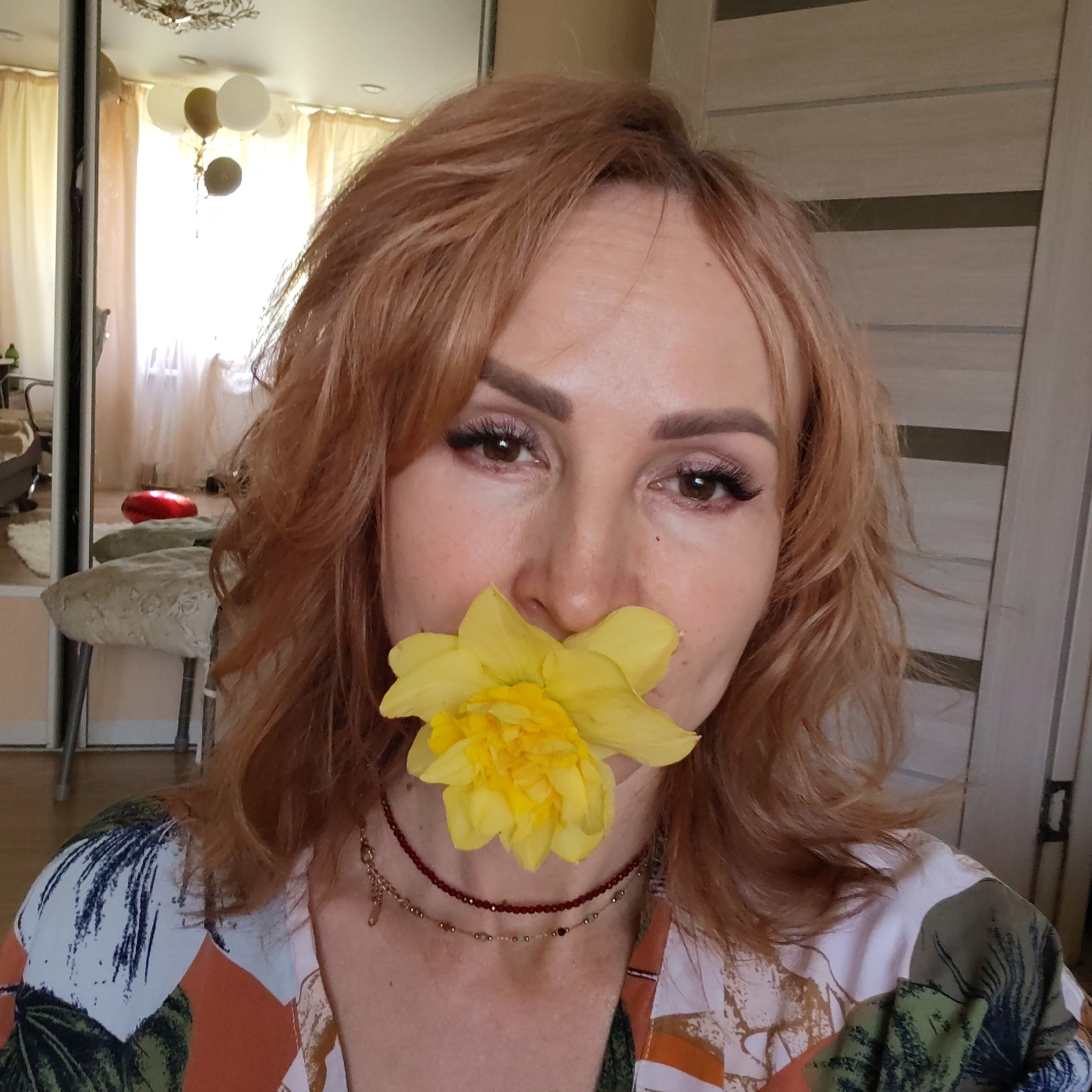 A middle-aged woman is the main subject of the image. She is holding a large yellow daffodil flower in her mouth, covering her lips and nose. Her hair is shoulder-length and reddish-blonde. She has brown eyes and wears makeup, including eyeshadow and false eyelashes. Around her neck are two delicate necklaces, one with red beads and the other with gold accents. She is wearing a patterned blouse with white, orange, and green designs.
The background is a room interior. A mirrored closet door is visible, reflecting a sitting area with stools and a red cushion. There are also balloons and a chandelier visible in the mirror's reflection, indicating a possible celebration. A beige door with horizontal panels is to the right of the woman.
The location is the city of Chaikovsky, Russia.