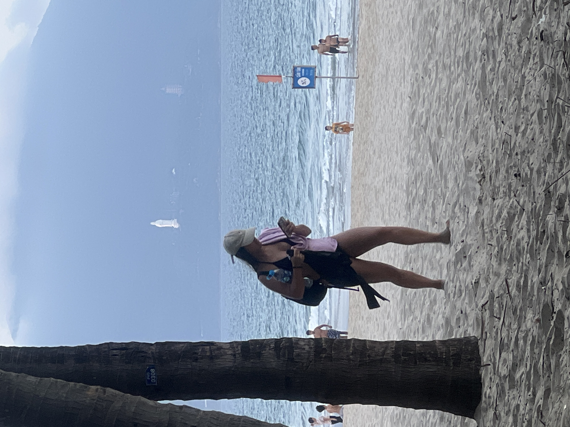 The image captures a woman on a beach in Da Nang, Vietnam, likely in the My Khe Beach area, given the presence of distant landmarks. She is walking on the sand, looking at her phone, and holding a water bottle and a pink towel. She is wearing a black swimsuit, a hat, and a backpack. In the background, the ocean stretches out, with waves gently rolling onto the shore. Several other people are visible on the beach and in the water, including children and men. In the distance, a white statue of a religious figure and a pagoda can be seen on a hazy mountainside, characteristic of the Son Tra Peninsula. The foreground is dominated by the textured sand and the trunks of palm trees, suggesting a tropical coastal setting. The overall atmosphere is relaxed and summery.