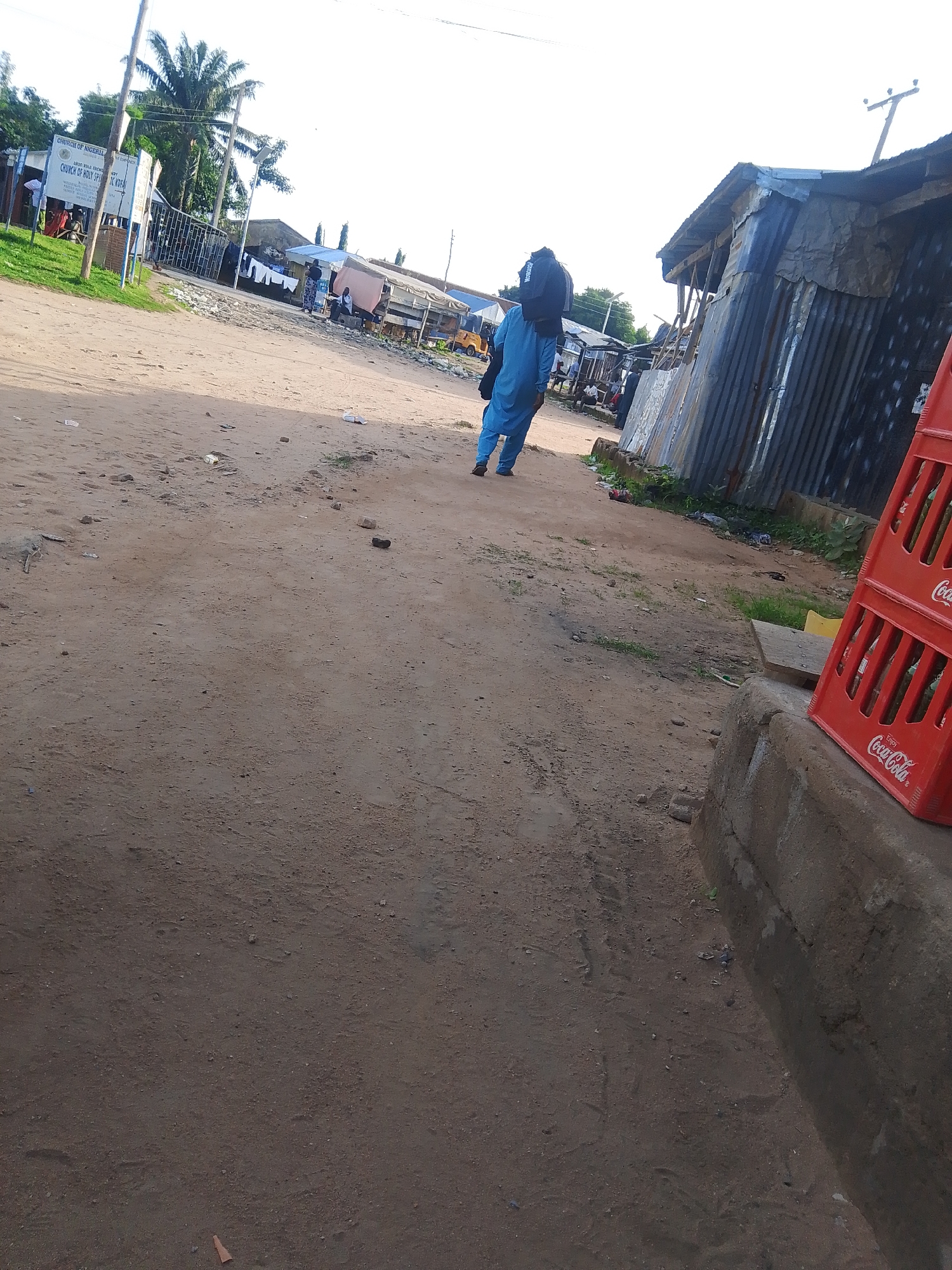 This media file depicts a scene in Jalingo, Nigeria, likely during the day with bright sunlight casting shadows, suggesting it's either morning or late afternoon. The setting is an unpaved, dusty road in what appears to be a market or a roadside settlement. In the foreground, the dirt road is scattered with debris, including small rocks and bits of trash.
The central subject is a person dressed in a blue kaftan, carrying a black backpack over their shoulders. They are walking away from the camera, down the road, towards a cluster of buildings. To the left of the road, there's a signpost with text that is partially legible. It reads "CHURCH OF NIGERIA" and "CHURCH OF HOLY SP..." suggesting a religious institution nearby. Further back, there are more people, some appearing to be engaged in commerce or daily activities near stalls and small shops constructed with corrugated metal.
To the right, a stack of red Coca-Cola crates is visible, partially obscuring a building made of corrugated metal. The overall atmosphere seems to be one of everyday life in a rural or semi-urban African setting. The sky is bright, and there are palm trees and other vegetation visible in the background, indicating a tropical or subtropical climate. The unpaved road and the makeshift structures suggest a less developed area. No strong emotions are discernible from the distant figures.