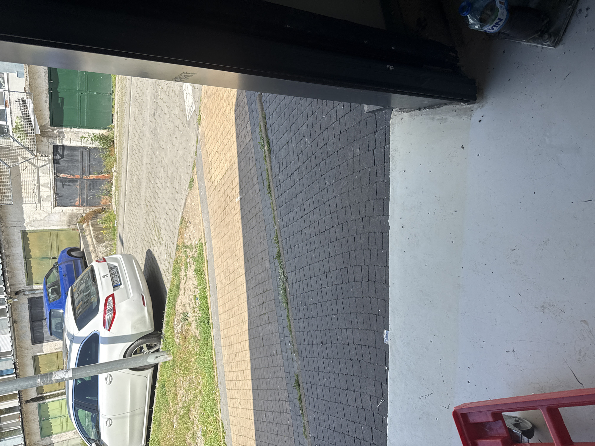 The image depicts a street scene in Stara Zagora, Bulgaria, observed from an indoor perspective through a doorway or window frame. A white station wagon and a blue hatchback are parked parallel to each other along the street's edge. The street is paved with gray and yellow interlocking concrete blocks. The buildings along the street are a mix of residential and garage structures. Some of the garage doors are painted green, while others are weathered and dark. A metal staircase is visible on one of the buildings. The immediate foreground of the image consists of a white concrete floor and a portion of a red trolley. A plastic water bottle rests near the upper edge of the doorway.