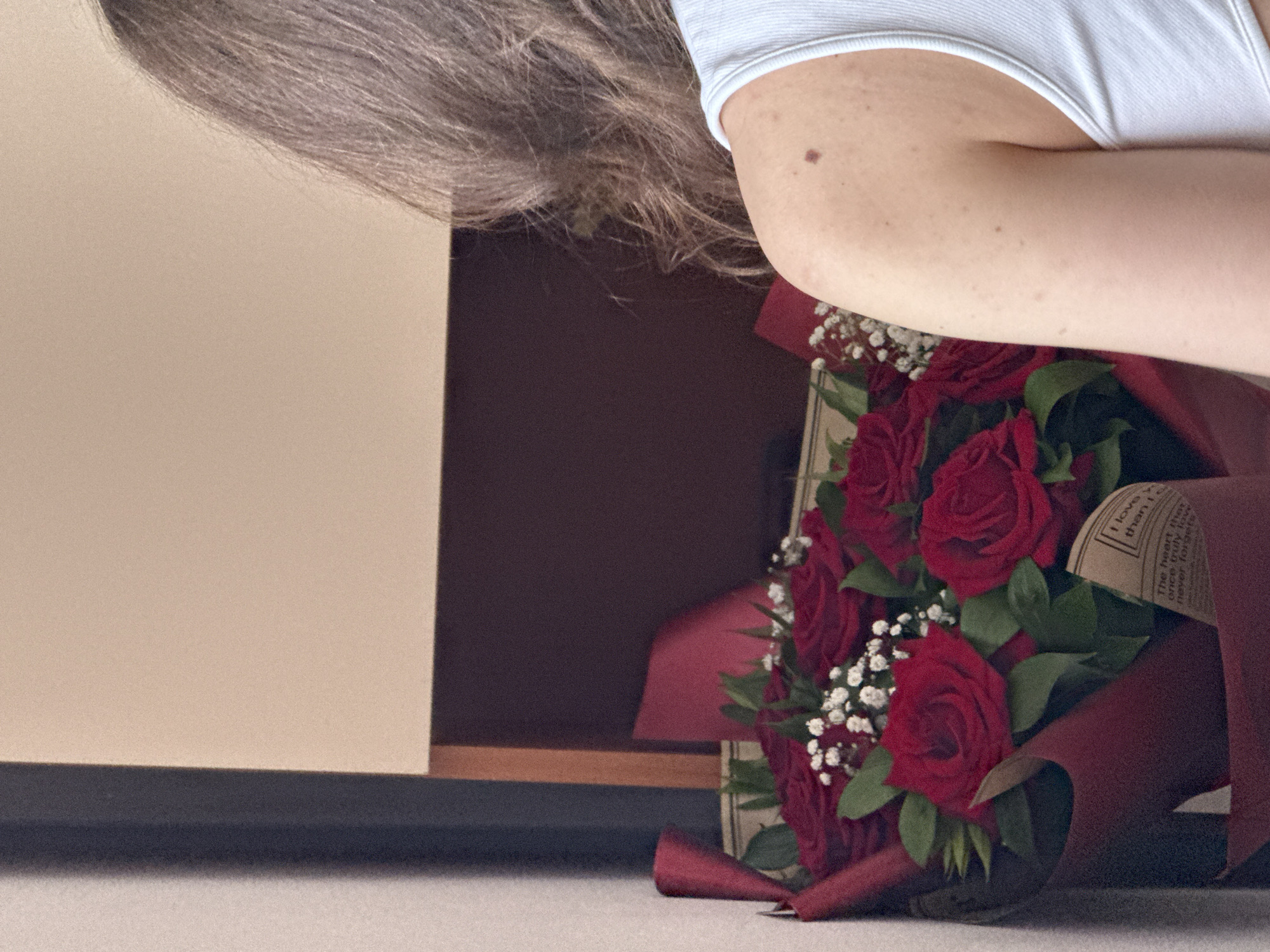 The image captures a close-up, top-down view of a person with a bouquet of red roses, taken in Varna, Bulgaria. The person's head and shoulder are visible in the upper left, with long, brownish-grey hair falling towards the left. A fair-skinned arm, marked with freckles and a distinct mole, extends from the upper right, wearing a white ribbed top.
The focus of the image is a vibrant bouquet of deep red roses, interspersed with small white filler flowers (likely baby's breath) and green foliage. The bouquet is lavishly wrapped in dark red or burgundy paper/fabric, which has a textured appearance. A small kraft paper tag or insert is visible within the wrapping, bearing partial text. The discernible phrases include "I know..." and "The heart that once truly loves never forgets."
The bouquet appears to be resting on a light grey or off-white surface in the lower right corner of the frame. The background behind the person and to the left consists of a plain, light beige wall, which meets a darker, possibly maroon or brown, background element or piece of furniture further to the right, creating a distinct horizontal line. The lighting suggests an indoor setting, likely during the day or under bright artificial light. The overall impression is one of a thoughtful gesture or gift.