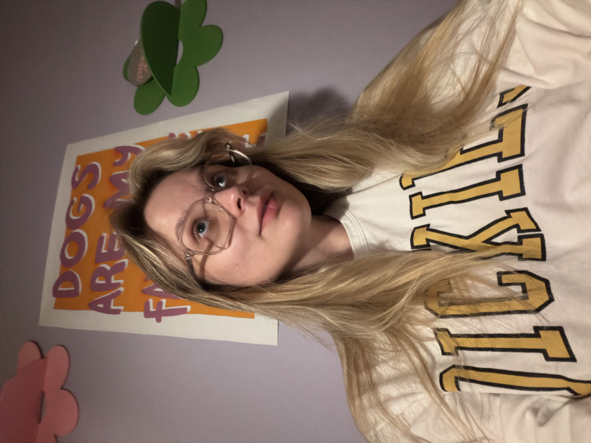 A young woman with long blonde hair is pictured indoors, possibly in a bedroom. She is wearing glasses and hoop earrings, and a white t-shirt with bold yellow and black lettering. The room is decorated with flower-shaped decorations and a framed poster with the words "DOGS ARE FAMILY" on it. The walls are a light purple color. There is no indication of this image being in Tel-Aviv, Israel.