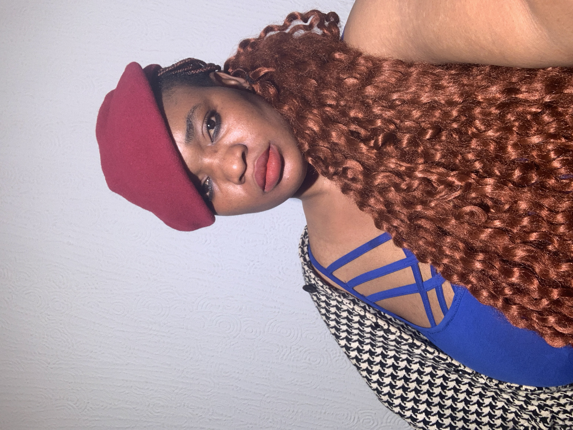 This is an indoor portrait of a young woman. She has dark skin and is looking directly at the camera with a composed, slightly contemplative expression. She wears a deep red beret tilted to her right side. Her voluminous, reddish-brown curly hair cascades over her left shoulder and chest. She has a reddish-orange lip color and noticeable eye makeup. Her attire includes a royal blue top or dress featuring intricate, multi-strap detailing on the right side of her upper back and shoulder. Over this, she wears an outer garment, possibly a jacket or shawl, with a distinctive black and white houndstooth pattern. The background is a light-colored (white or very light grey) wall with a subtle, embossed circular pattern. The image is tilted significantly, giving the impression that the subject is leaning or lying down for the photograph. The lighting is soft and even, characteristic of indoor artificial light. No text is visible in the image. The activity is posing for a portrait photograph.