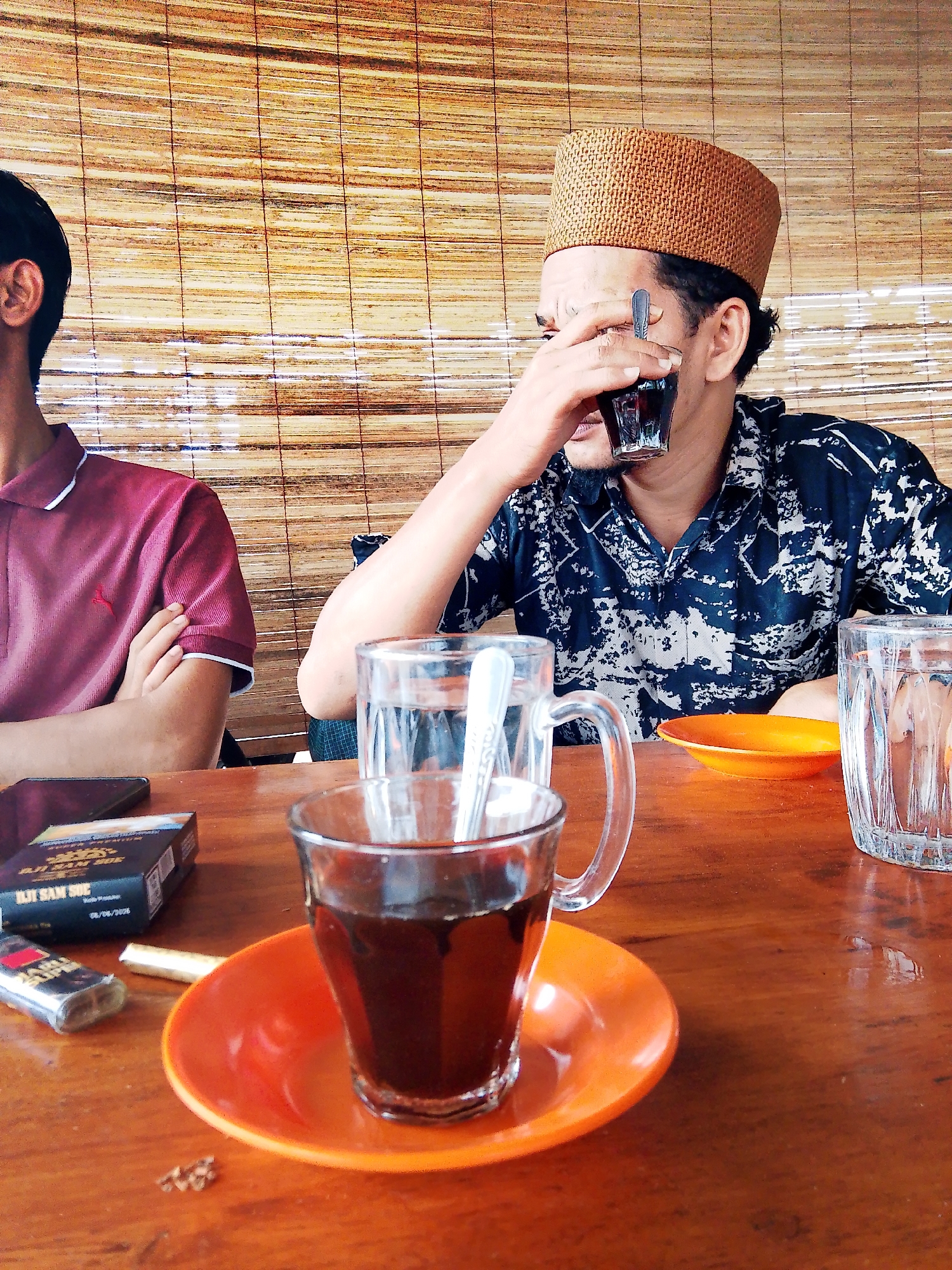 In Keudejungkagajah, Indonesia, two men sit at a wooden table, their faces partially obscured by a woven bamboo screen. The man on the right, wearing a tan kufi and a dark patterned shirt, raises a glass of dark liquid to his face with his right hand. In front of him, a small orange saucer holds a glass of the same dark liquid, a spoon visible inside. To his left, a man in a maroon polo shirt with white trim sits with his arms crossed. A pack of cigarettes and a lighter are on the table in front of him. Another glass of water is visible on the far right of the table. The lighting suggests an indoor or shaded outdoor setting.