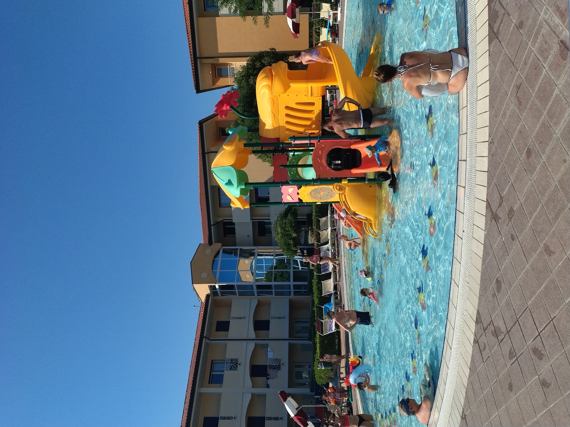 The image depicts a bright, sunny day at a resort pool in Caorle, Italy, with a clear blue sky overhead. The pool area features a vibrant yellow and orange water playground with slides and climbing structures, designed for children. Several individuals, including adults and children, are enjoying the water. Some are swimming or wading, while others are interacting with the playground equipment. In the background, modern hotel buildings with orange-tiled roofs are visible, suggesting a tourist accommodation. Lounge chairs are arranged around the pool, and some are occupied by people relaxing. The pool itself is a clear turquoise blue, with decorative fish shapes visible on the bottom. The scene is one of leisure and recreation, typical of a summer vacation destination.
