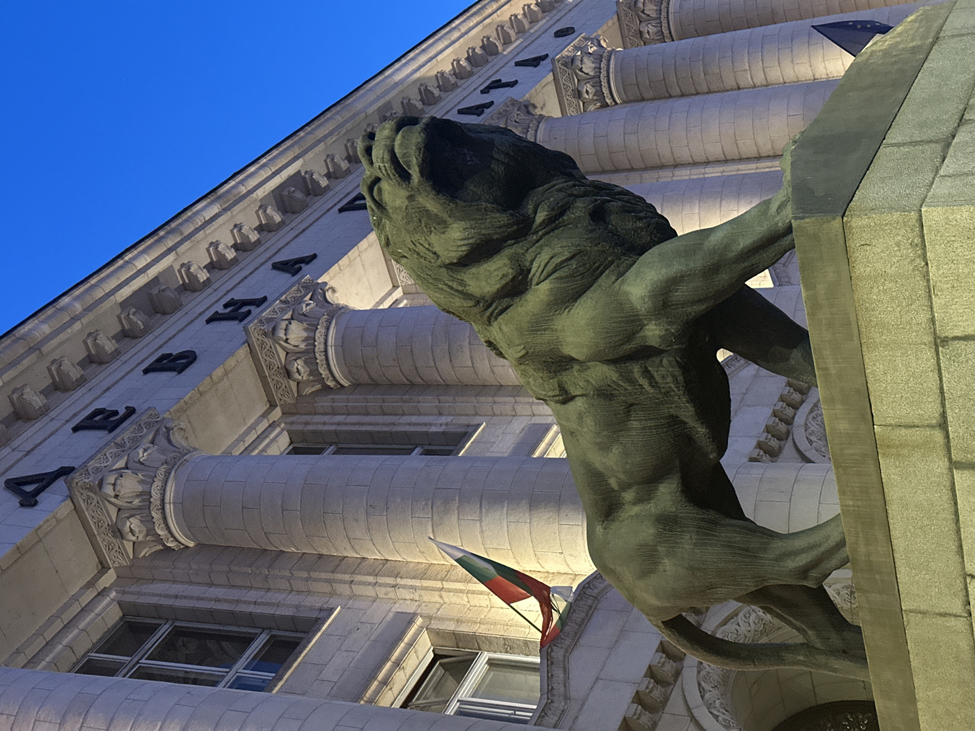 The image captures a low-angle view of a building facade in Sofia, Bulgaria, featuring a prominent lion sculpture. The building's architecture is characterized by ornate details, including decorative moldings, fluted columns, and recessed windows. A Bulgarian flag is visible hanging from one of the window bays. The lion sculpture, made of a dark metal material, is positioned on the building's exterior, adding a symbolic and artistic element to the structure. The sky above is a clear, deep blue. The building has letters inscribed on its face that appear to spell out "ДЪРЖАВНА".