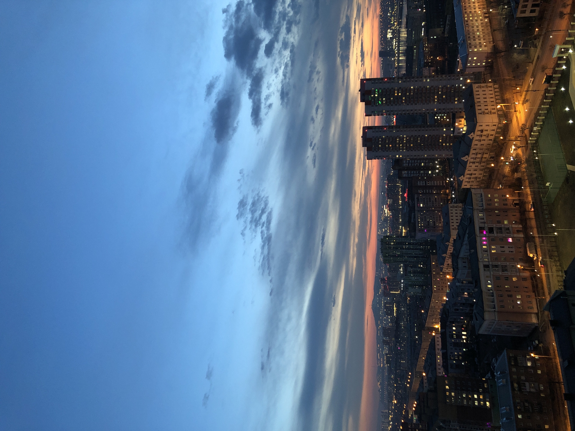 An expansive aerial or high-vantage point view captures the city of Krasnoyarsk, Russia, at dusk or twilight. The scene is dominated by a vast sky that transitions from a deep, clear blue at the top-left to lighter blues and greys, then to vibrant orange and pink hues along the horizon on the right, indicative of a sunset. Scattered clouds, varying from dark grey and dense to wispy and light, are distributed across the sky.
Below the dramatic sky, the urban landscape stretches into the distance. Numerous multi-story buildings, likely a mix of residential and commercial structures, populate the city. Many of these buildings have illuminated windows, casting a warm glow across the scene as evening sets in. Streets and thoroughfares are visible, lined with glowing streetlights that create intricate patterns of light throughout the city. A faint, distant outline of hills or mountains can be observed along the horizon, blending with the sunset colors. The overall impression is that of a bustling city transitioning into the night, viewed under a vast and colorful sky. No specific human activity or discernible text is visible within the frame.