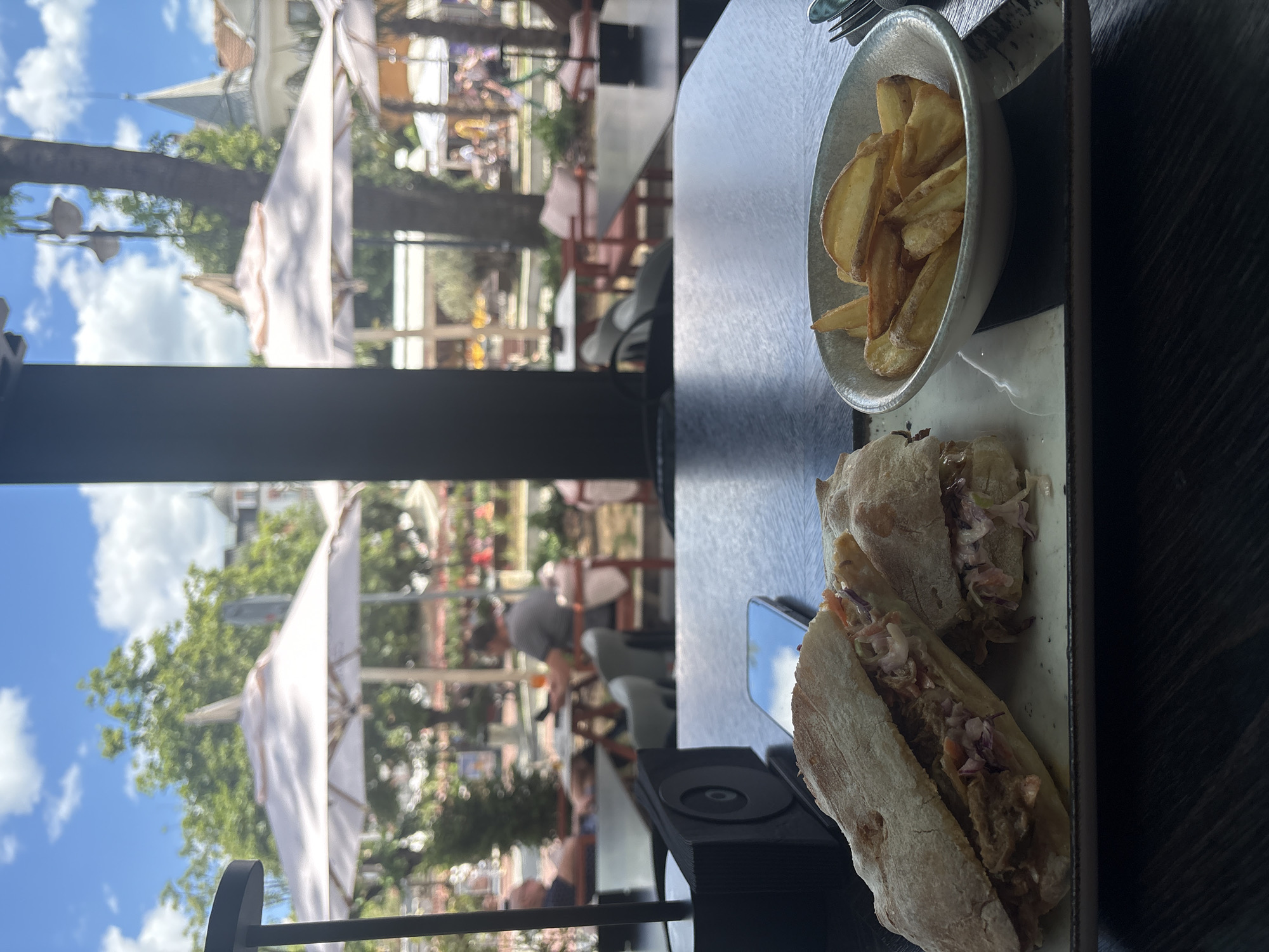 In the foreground, on a dark wooden table, is a meal served on a rectangular plate. The meal consists of two halves of a sandwich on ciabatta bread, filled with pulled meat and coleslaw, and a side of thick-cut potato wedges served in a light-colored bowl. A smartphone is visible on the table next to the food. To the left, a black object, possibly a napkin holder or speaker, is also present. The background, viewed through a large window or glass partition, shows an outdoor seating area with tables, chairs, and umbrellas, suggesting a cafe or restaurant with an outdoor patio. People are visible in the distance, some seated at tables and others walking. Trees, buildings with architectural details, and a bright blue sky with scattered clouds complete the scene.