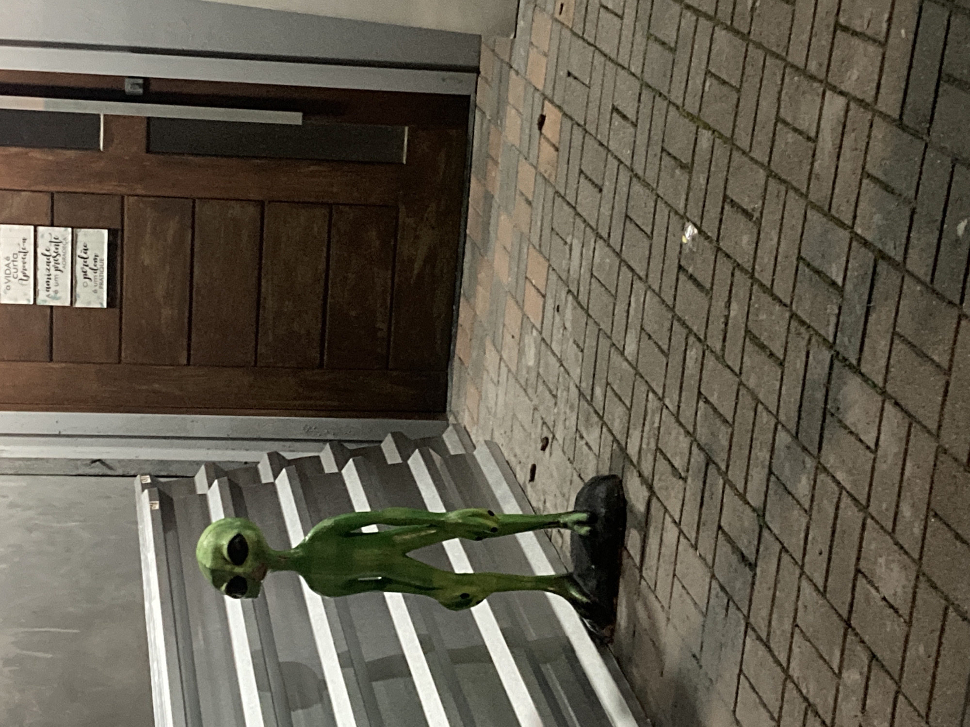 A green, humanoid alien figure stands on a black base near the base of some steps. The steps are white and are made of corrugated metal. Behind the steps, there is a wall that looks like it is concrete. Next to the wall and steps is a dark wood door with a metallic handle. On the door are a few white placards. The ground is comprised of grey brick pavers. The location is in Florianópolis, Brazil.