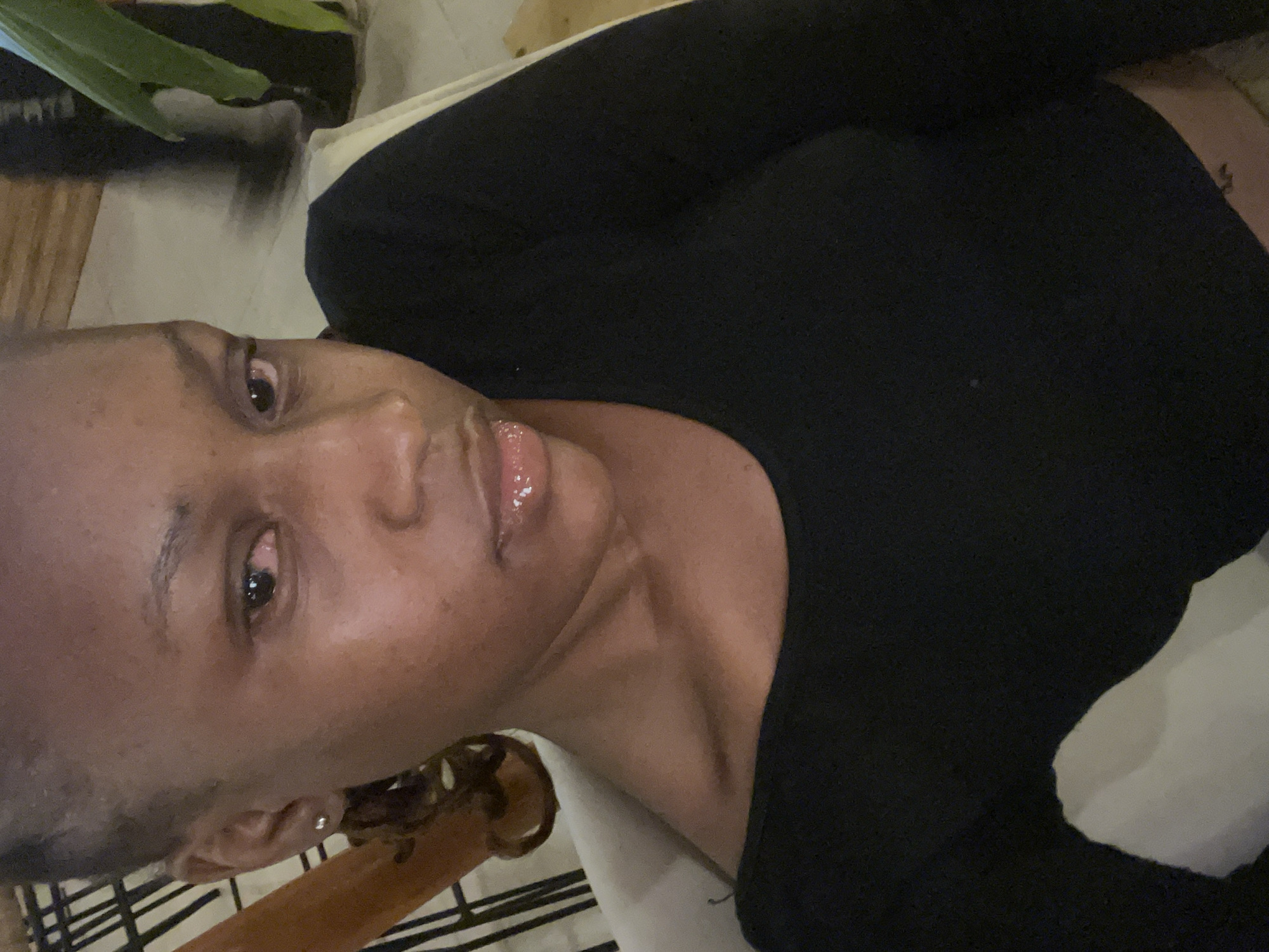 A young woman, with dark skin and braided hair, is captured in a close-up selfie. She is wearing a black, long-sleeved shirt. Her right eye appears red and irritated. Her lips are glossy. She is sitting on a light-colored surface, possibly a chair. Behind her, a blurred background shows vertical bars, suggesting a fence or grate, and a large green plant. The image appears to be taken indoors. There is no identifiable location in the city of Gura Top, Nigeria.