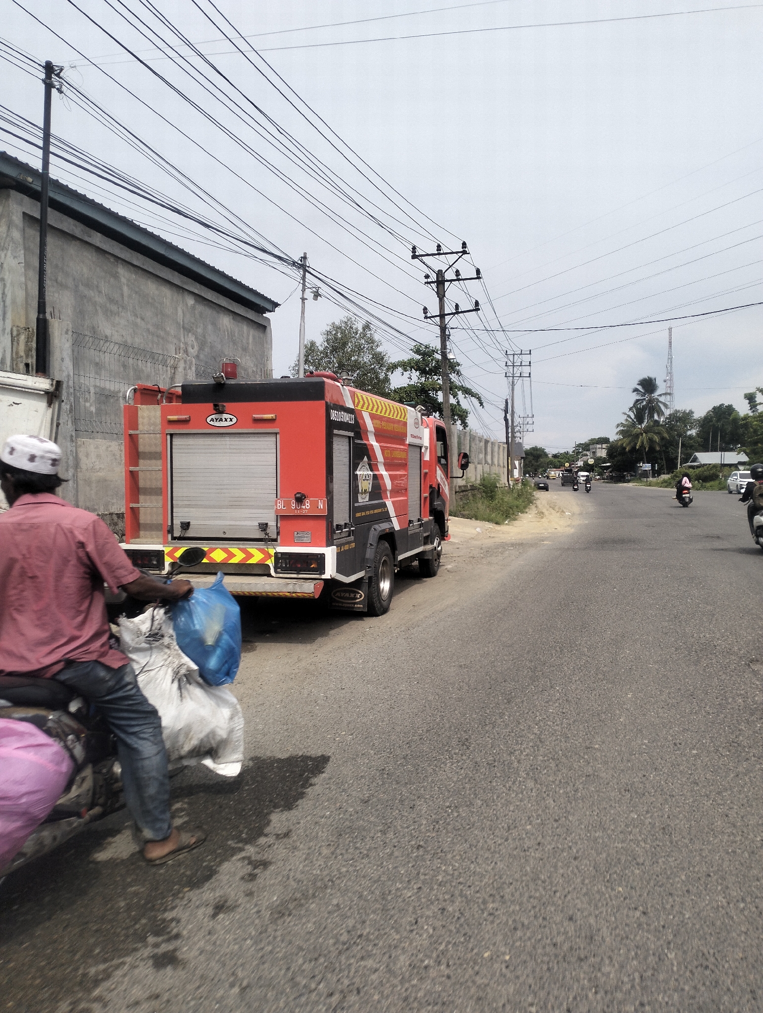 The image shows a street scene in an urban environment. A red fire truck is parked on the right side of the road, facing away from the viewer. It has "AYAX" written on its side in white lettering and a license plate that reads "BL 9048 N". A person wearing a red shirt and a white cap is riding a motorcycle in front of the fire truck, carrying large bags. The road is paved and has traffic lines. In the background, there are buildings, trees, power lines, and a cell tower. The sky is overcast.