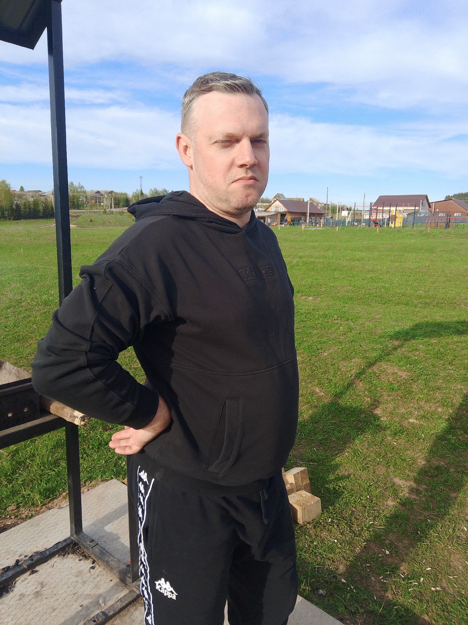 A man stands outside in Фоки, Russia. He is dressed in a black Kappa hooded sweatshirt and matching sweatpants. He is leaning against a dark metal structure, possibly a barbecue grill. In the background is a green field with a few houses and what appears to be a playground in the distance. The sky is blue with scattered clouds.