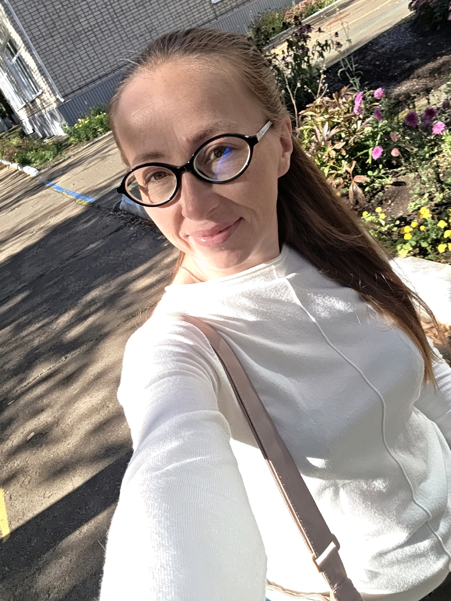 A woman with brown hair, wearing black-rimmed glasses and a white long-sleeved shirt, stands outside, taking a selfie. The background shows a building with a grey brick facade, a sidewalk with yellow and blue markings, and a flower bed with pink and yellow blossoms. Sunlight casts shadows across the ground, indicating it is daytime. The woman is carrying a purse or bag with a tan strap slung across her body. The image appears to have been taken on a street or walkway in Chaikovsky, Russia, likely in a park or residential area given the presence of flowers and a building.