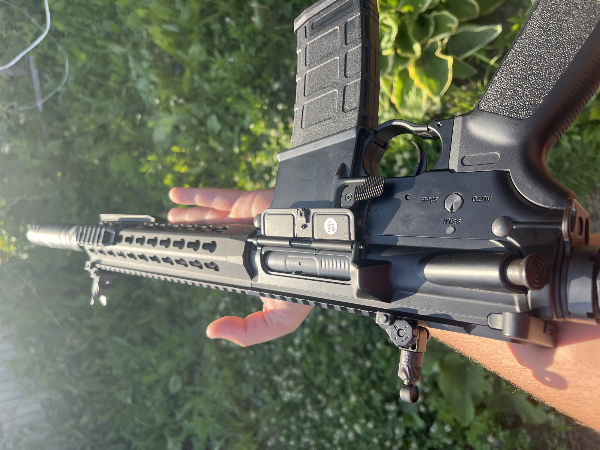 A close-up, low-angle shot shows a person holding a black semi-automatic rifle outdoors. The rifle is held in the right hand, with fingers gripping the textured pistol grip and the thumb resting on the selector switch. The rifle is pointed away from the viewer and angled upwards, with the barrel extending out of frame at the top. The receiver of the rifle is visible, featuring a magazine release lever, bolt catch, and safety selector with markings for "SAFE", "SEMI", and "AUTO". A black rifle magazine is inserted into the mag well. The background consists of lush green foliage, suggesting a natural outdoor setting. There is no visible indication of the city of Vasilyevo, Russia.