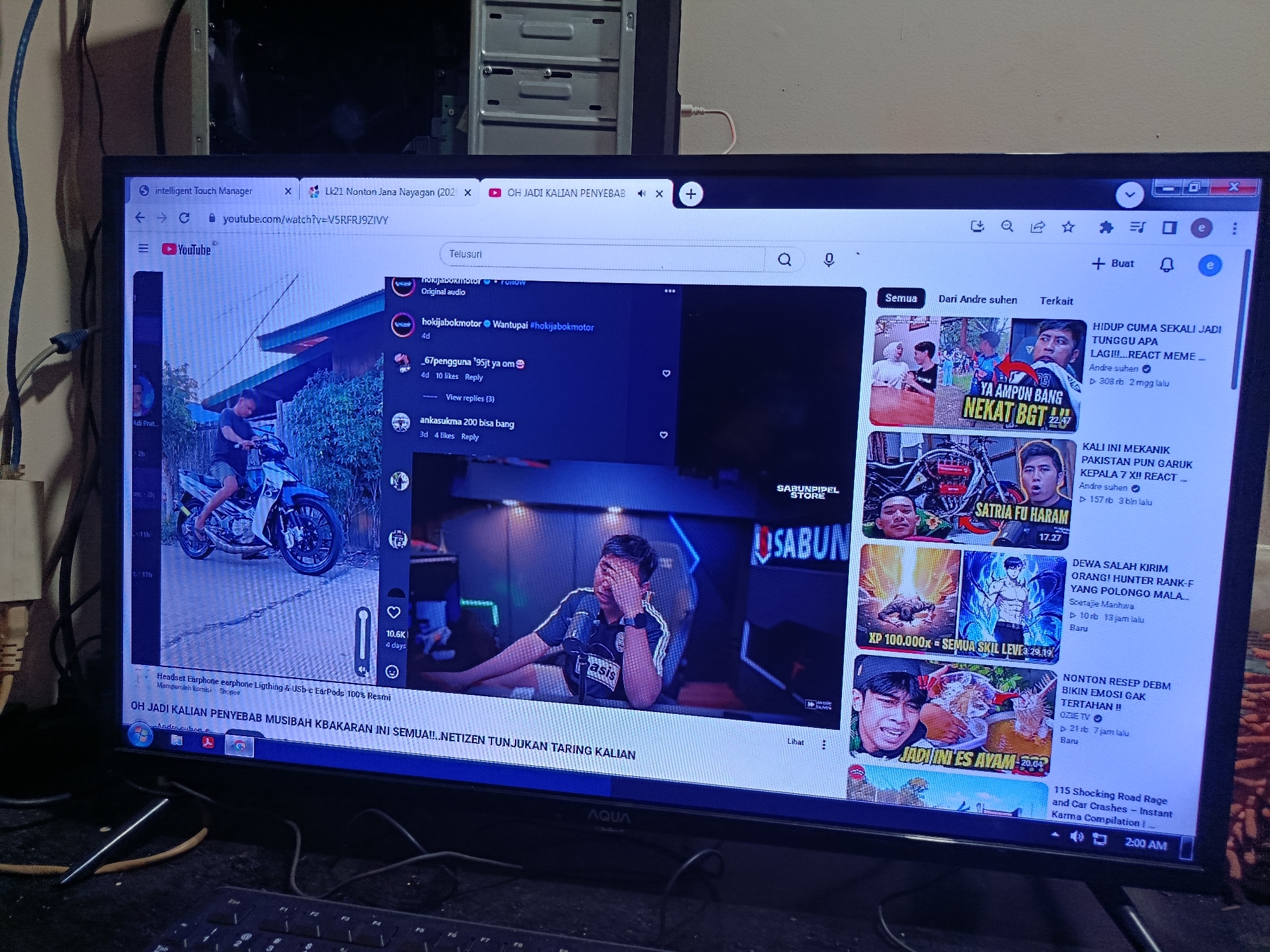 The image shows a computer monitor displaying a YouTube page. The page appears to be open in a web browser, with the URL bar visible at the top. The YouTube video playing shows a man on a motorcycle. There are also comments visible on the page, with user names and reply options. The monitor is placed on a desk, with a keyboard visible in the foreground. The background is blurred, but appears to be an indoor setting.