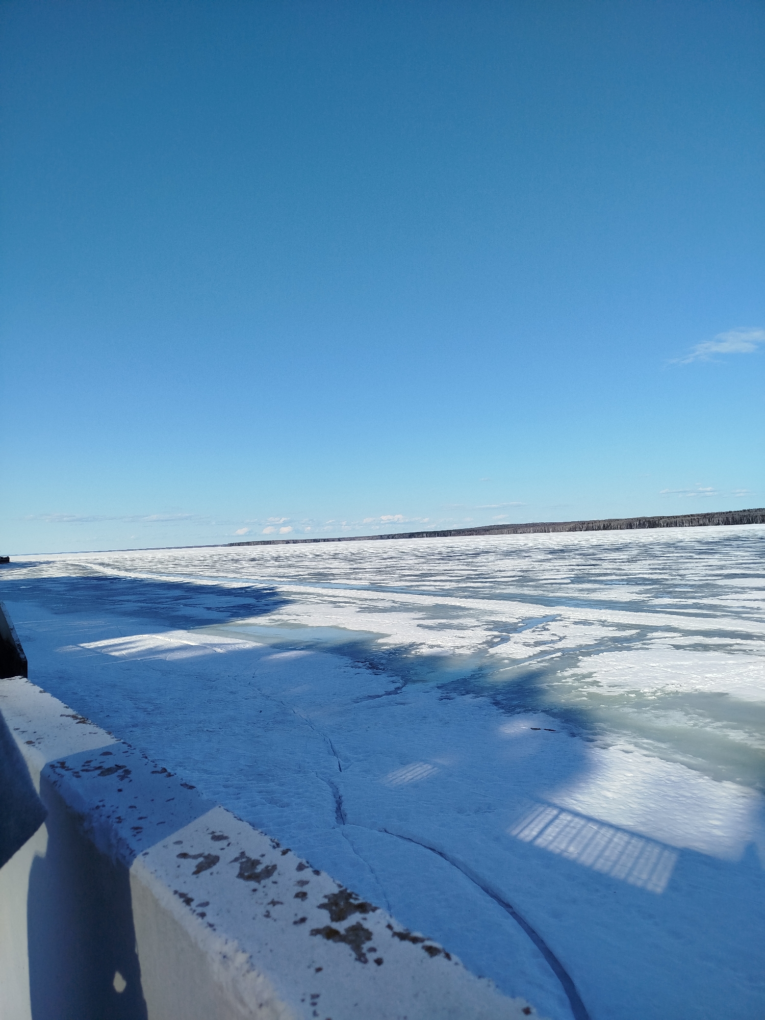 The image depicts a wide, frozen expanse of water, likely a river or lake, under a clear, bright blue sky with sparse clouds. The foreground includes a concrete barrier, possibly a bridge or embankment, casting shadows onto the ice-covered surface. The ice appears broken and patchy, with areas of open water visible. In the distance, a line of trees is visible along the horizon. The location is likely Chaikovsky, Russia, given the characteristics of the landscape. The image conveys a cold, wintry scene.