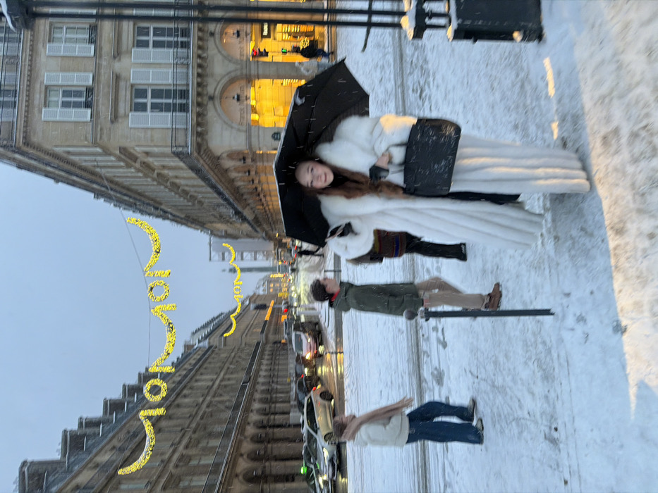A street scene depicts a woman in a white fur coat holding a black umbrella. Two people are walking behind her, one wearing a green coat and khaki pants, and another in a beige shirt and blue jeans. Buildings line the street, decorated with golden holiday lights. The ground is covered in snow. Cars and a taxi are parked on the side of the street.