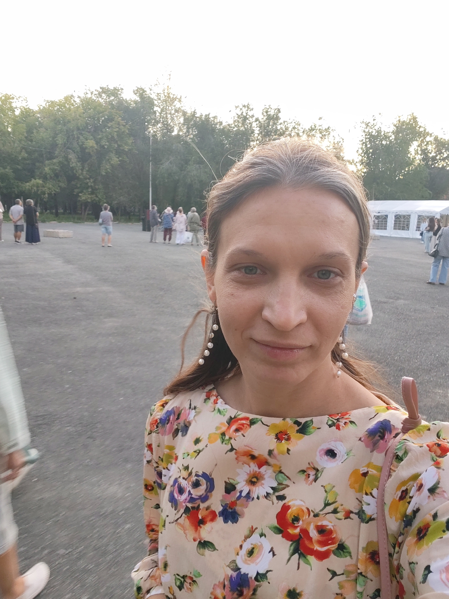 In an outdoor setting, a woman with light brown hair and blue-green eyes takes a selfie. She is wearing a light-colored, floral-patterned shirt with pearl earrings. Behind her, a group of people are standing in the distance, with trees and a white tent structure visible. The ground is paved, suggesting an outdoor event or gathering space.