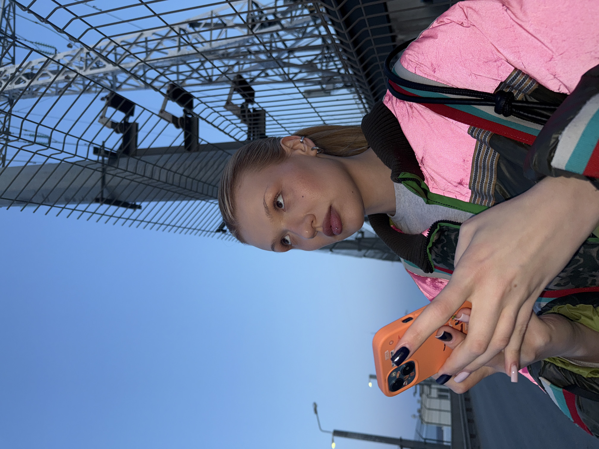 A young woman with blonde hair, wearing a pink jacket, is holding an orange smartphone. The woman is fair-skinned and has multiple earrings in her left ear. She appears to be looking at the phone screen. The background includes a metal structure, possibly part of an amusement park ride or industrial framework. The sky is a clear blue. Streetlights are visible in the lower portion of the image. The photograph was taken outdoors in the city of Chaikovsky, Russia.