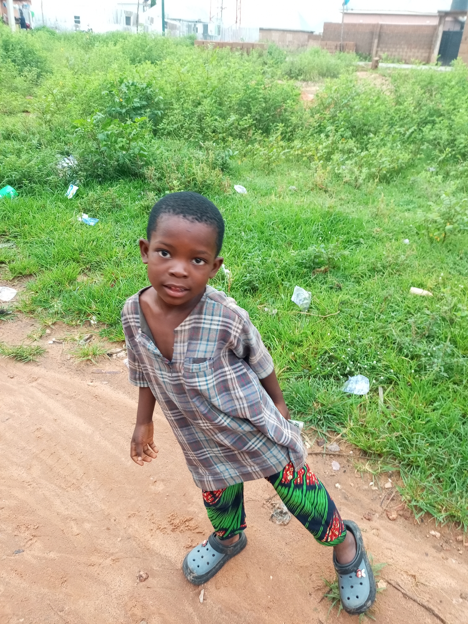A young Black boy, approximately 7 years old, stands on a dirt path in a somewhat overgrown, grassy area. He is wearing a plaid, collared shirt that is open at the neck and colorful green and red patterned pants. He has on grey Croc-style shoes. The background shows a mix of green grass, bushes, and some scattered debris, likely plastic bags and wrappers. In the distance, buildings under construction or in disrepair are visible. The overall scene suggests an outdoor environment in a developing area, possibly in Tempure, Nigeria, based on the context provided. The boy is looking directly at the camera with a neutral expression.